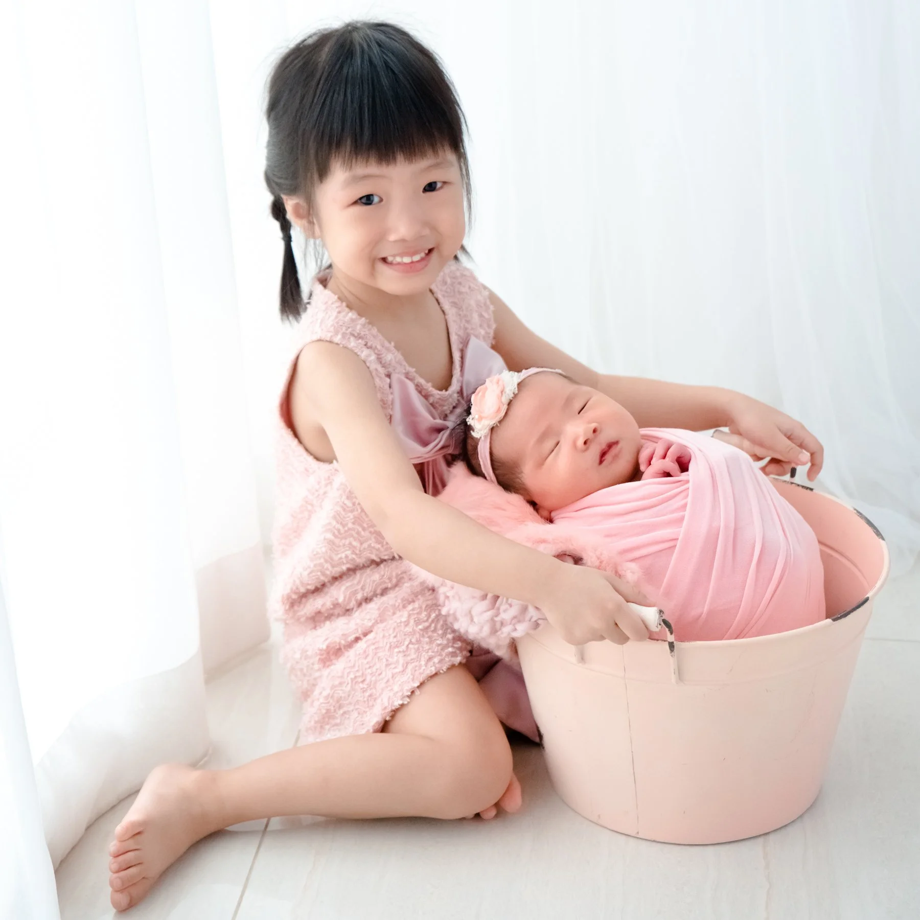 Older sister in pink sitting beside her newborn sibling wrapped in pink in a small basket