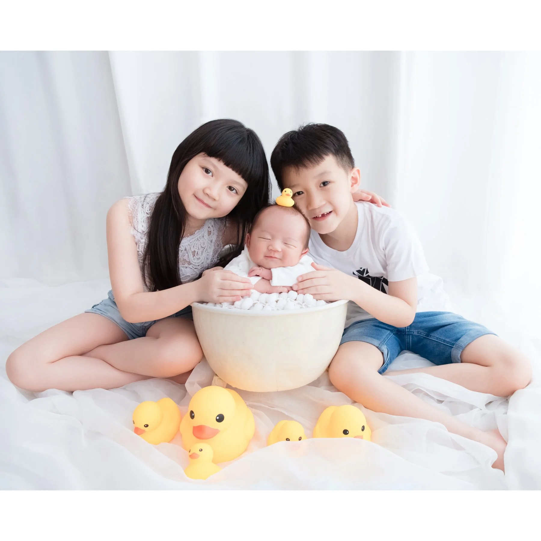 Older sister and brother cuddling their newborn sibling in a white bathtub with yellow rubber ducks