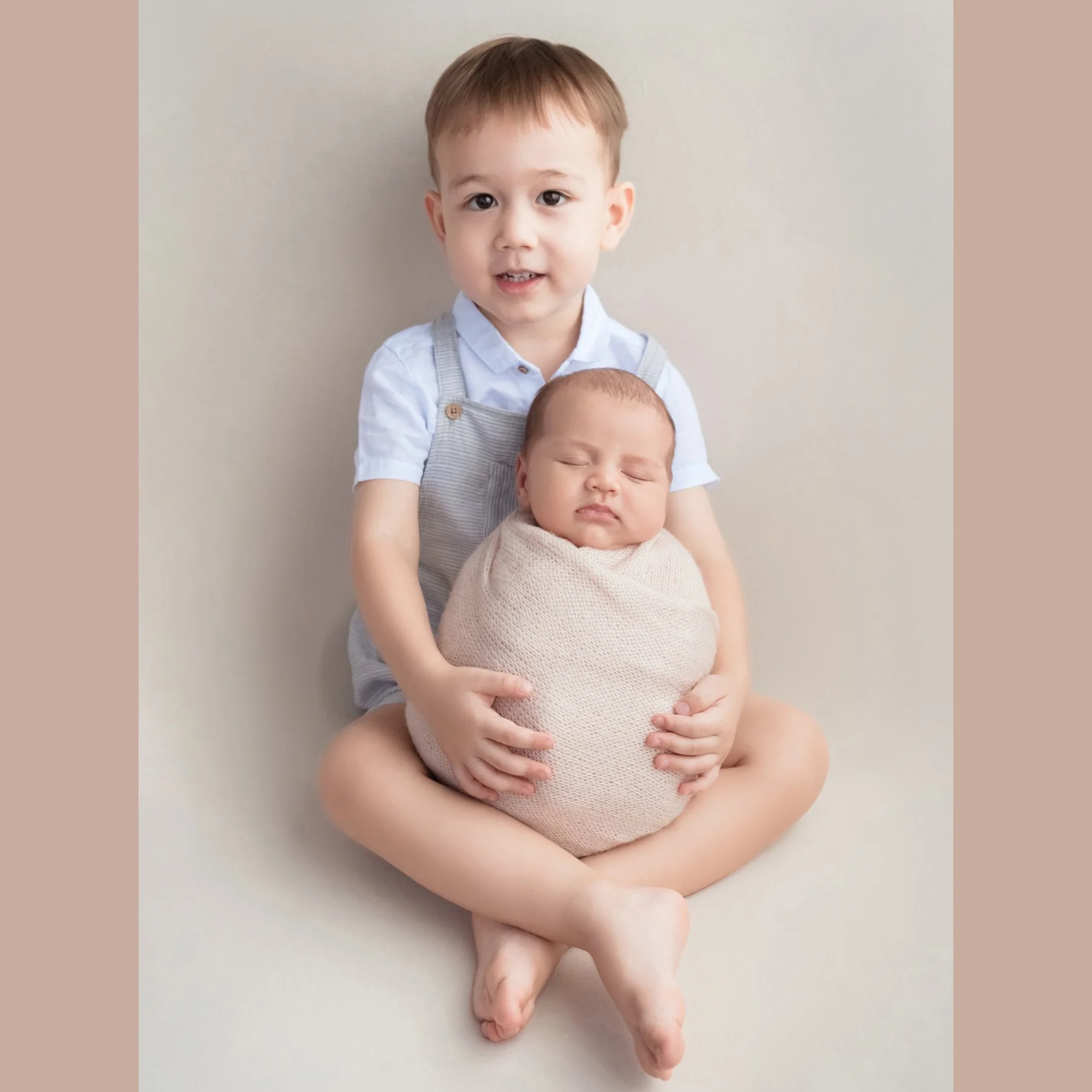 Older brother seated with his newborn sibling wrapped in cream against a neutral backdrop