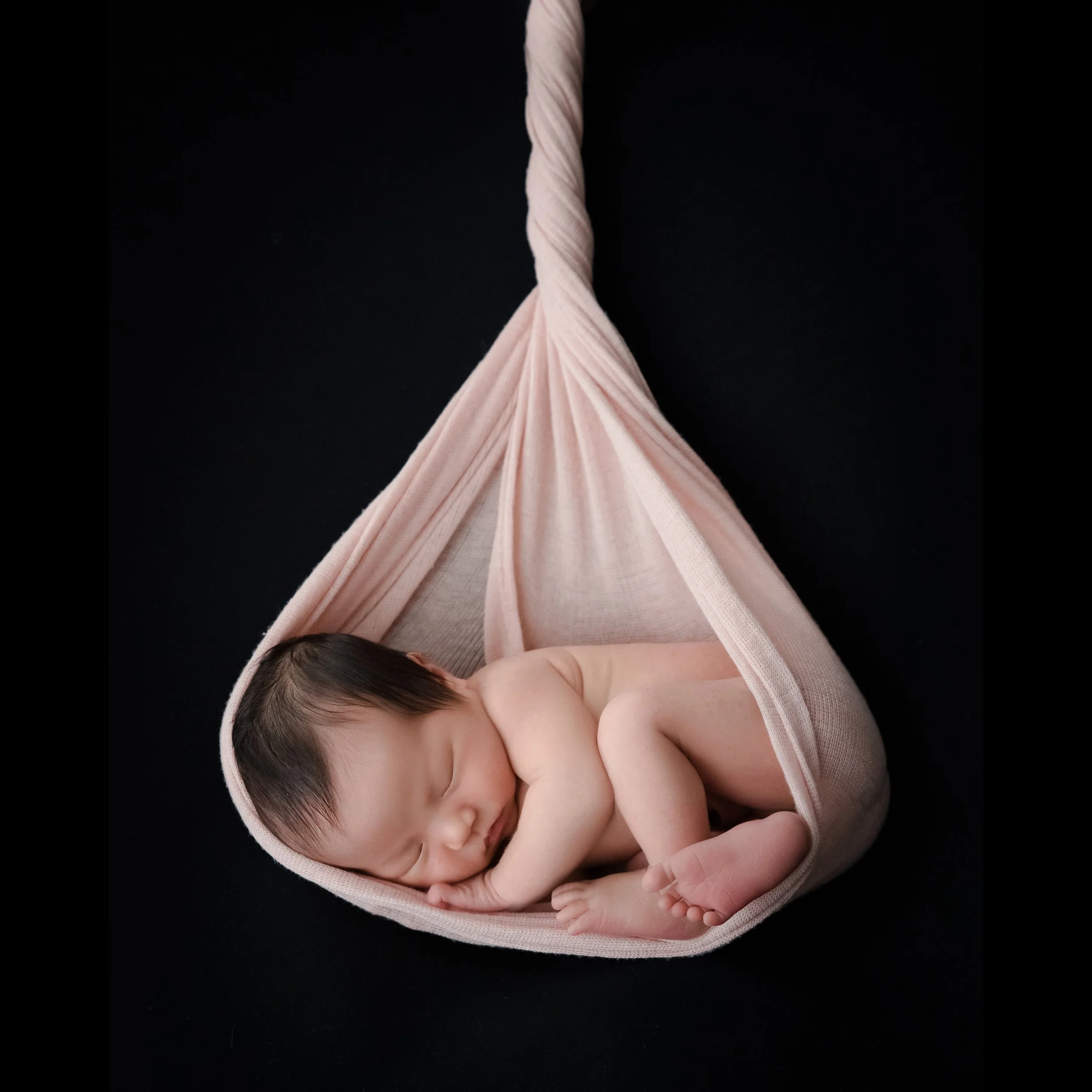 Sleeping newborn wearing a pearl headband curled in a pale pink fabric hammock against a black backdrop