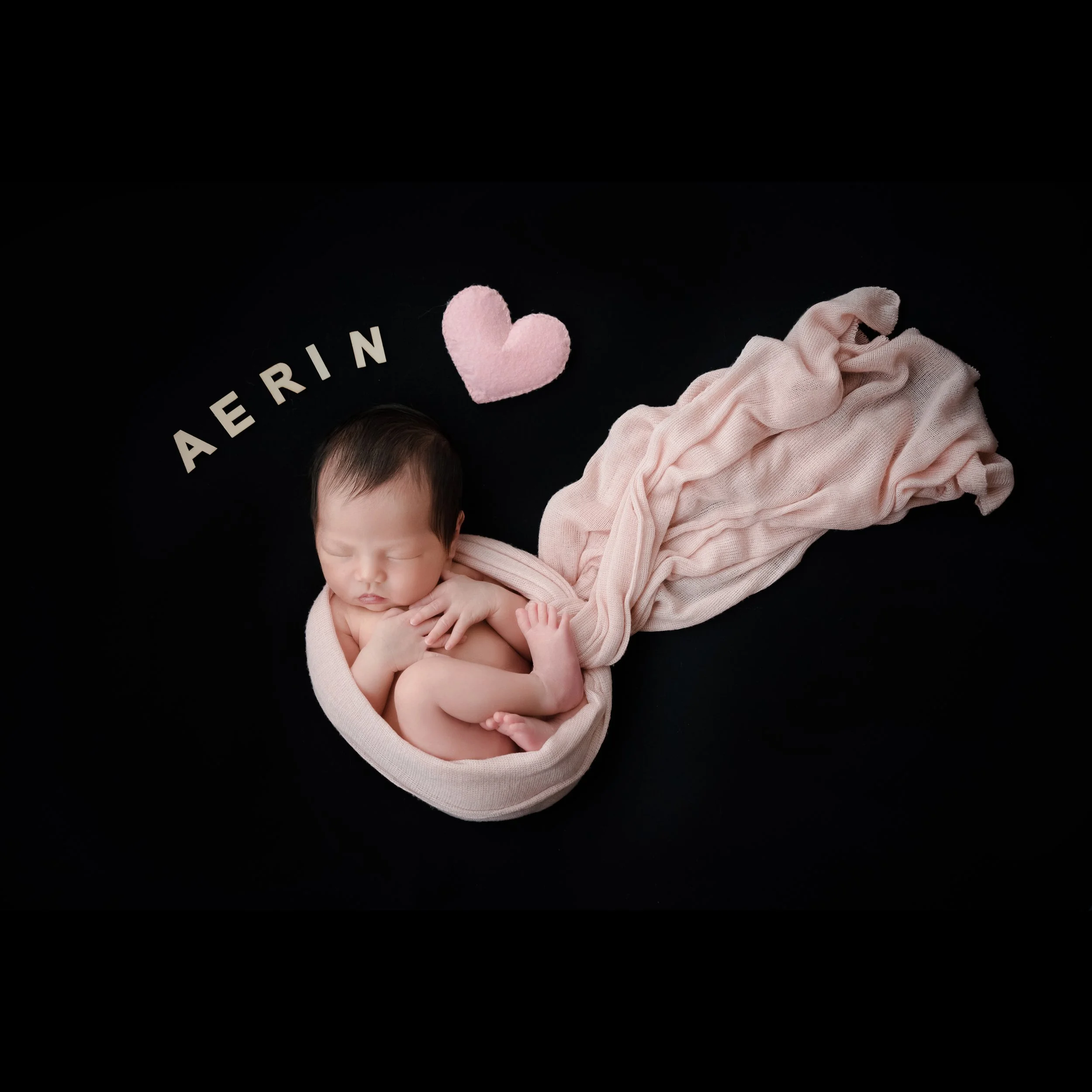 Sleeping newborn wearing a pearl headband curled in a pink fabric nest beside a pink heart decoration on a black backdrop