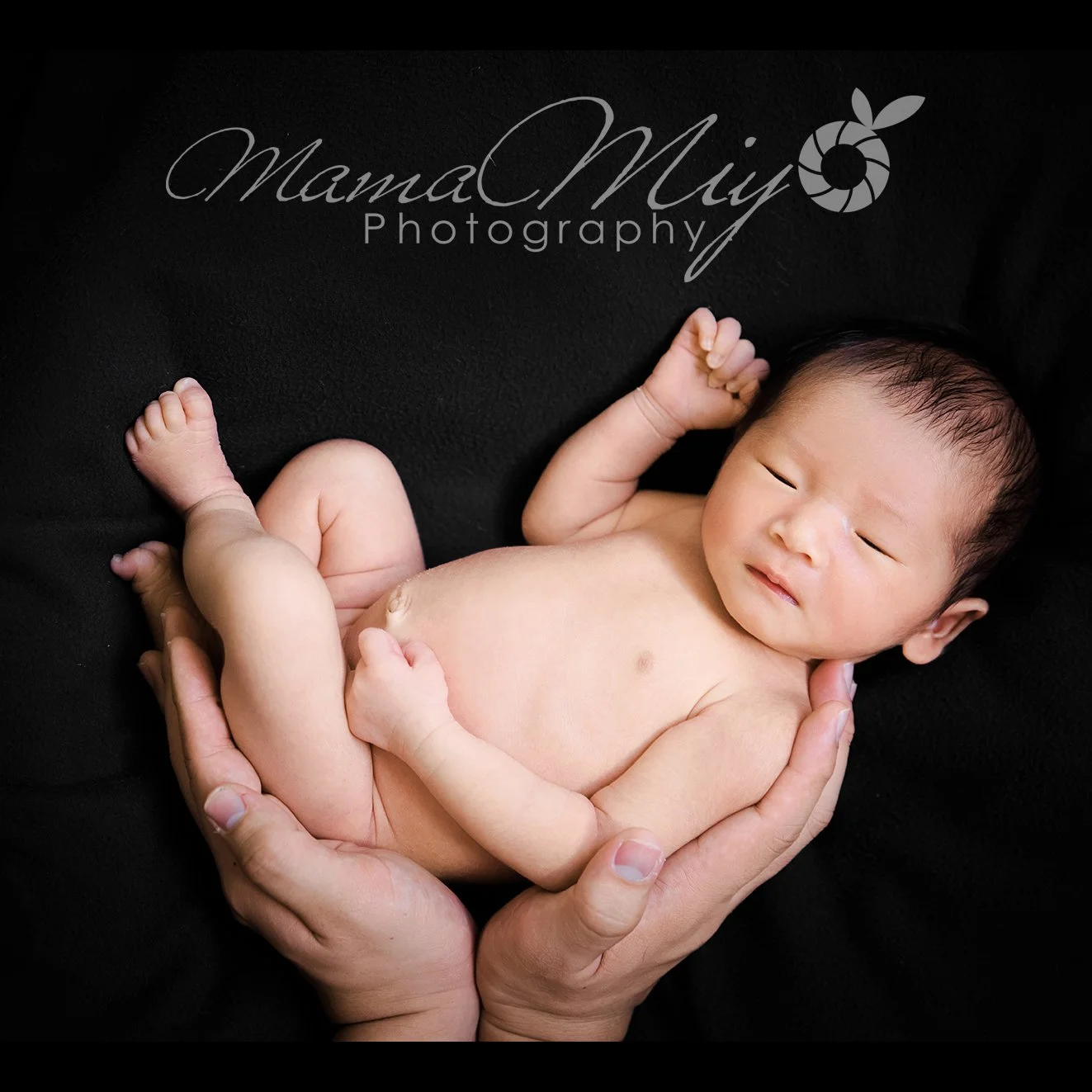 Smiling newborn with one arm raised and legs curled while supported by adult hands against a black backdrop