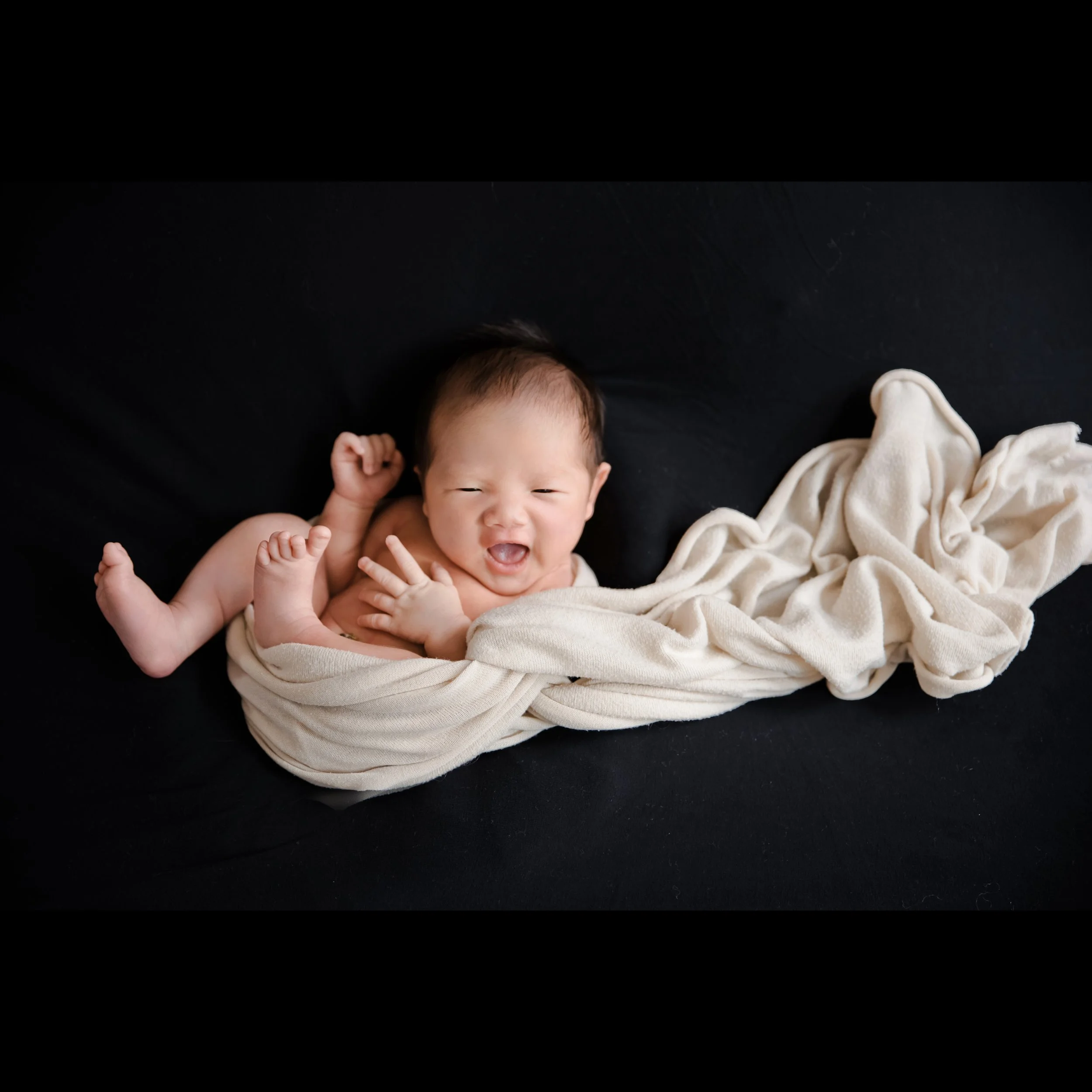 Yawning newborn loosely wrapped in cream fabric lying on a black backdrop with one foot raised