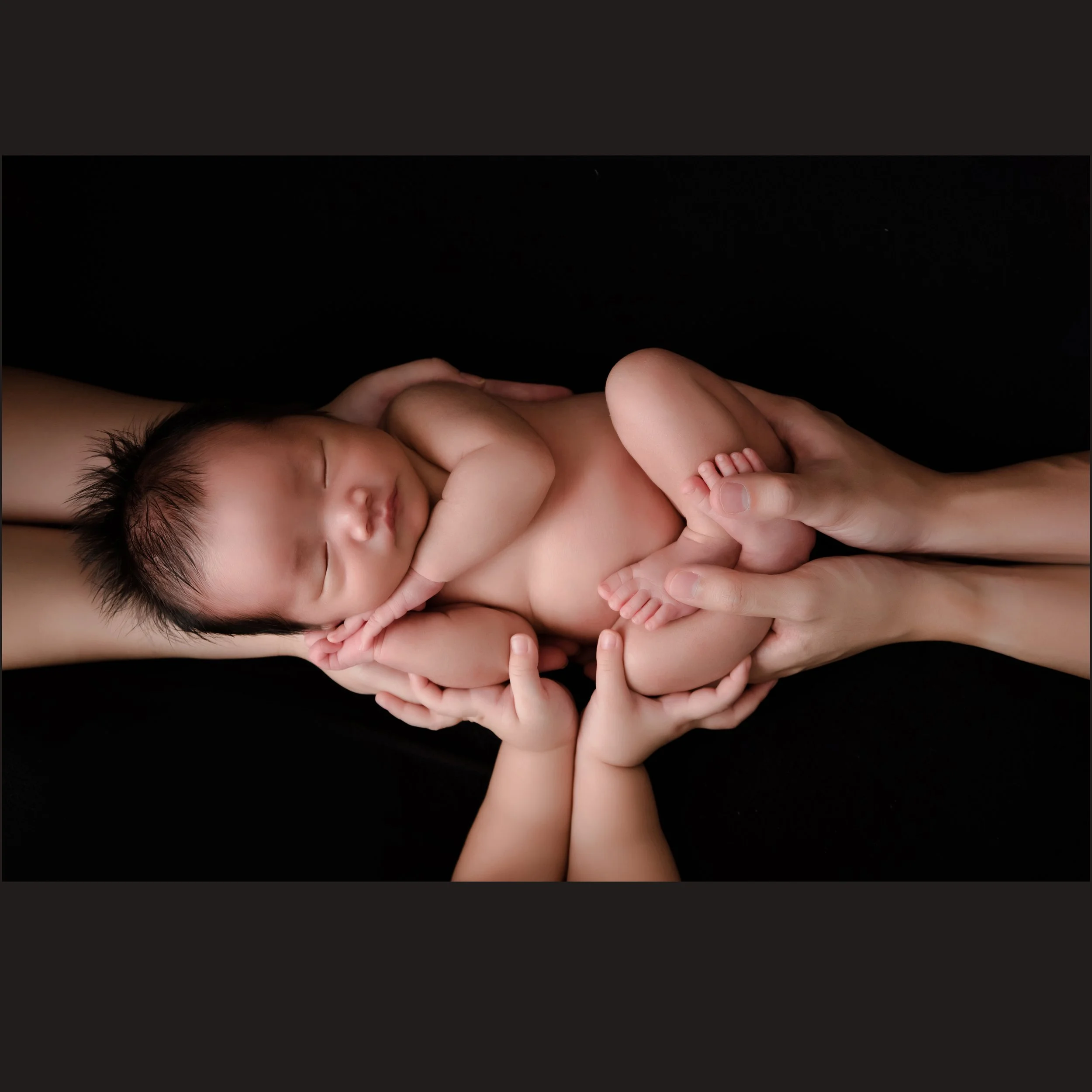 Sleeping newborn curled on one side while supported by several adult hands against a black backdrop