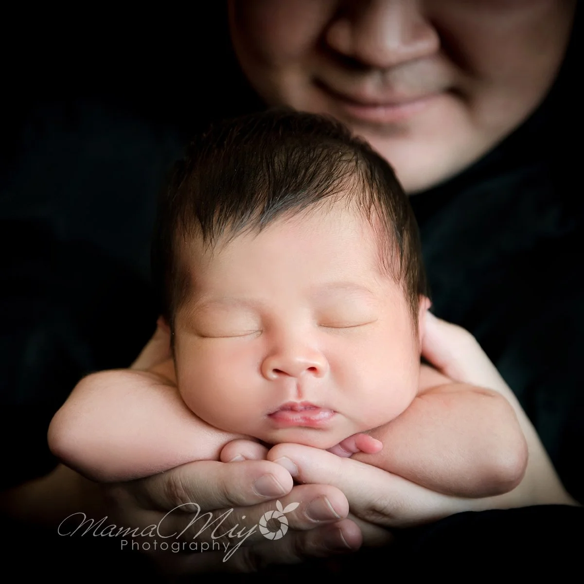 Close-up of a sleeping newborn resting the chin on folded arms in adult hands with an adult face gently leaning above against a black backdrop