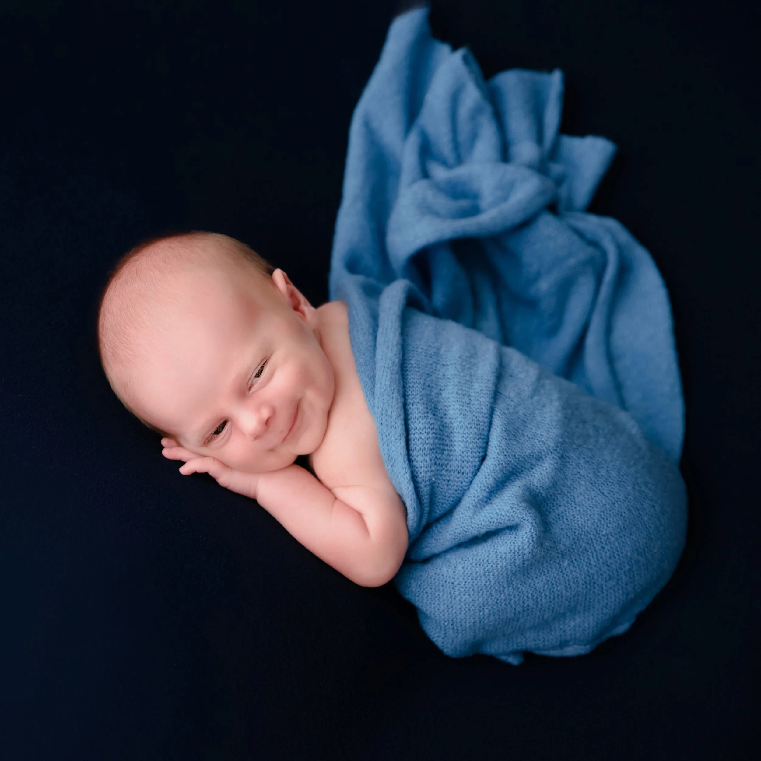 Awake newborn loosely wrapped in blue fabric lying on one side with hands beside the cheek against a black backdrop