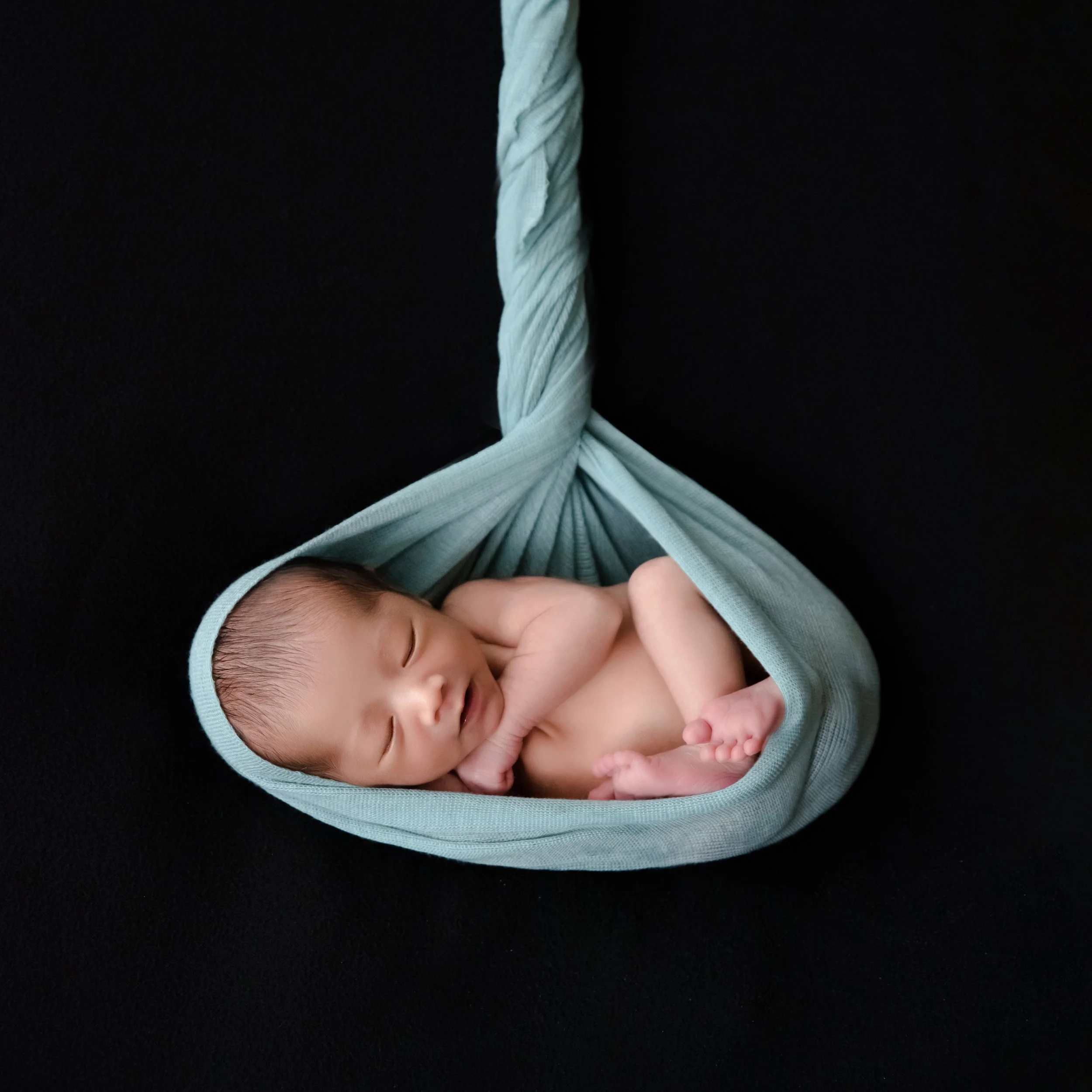 Sleeping newborn curled in a sage-green fabric hammock against a black backdrop