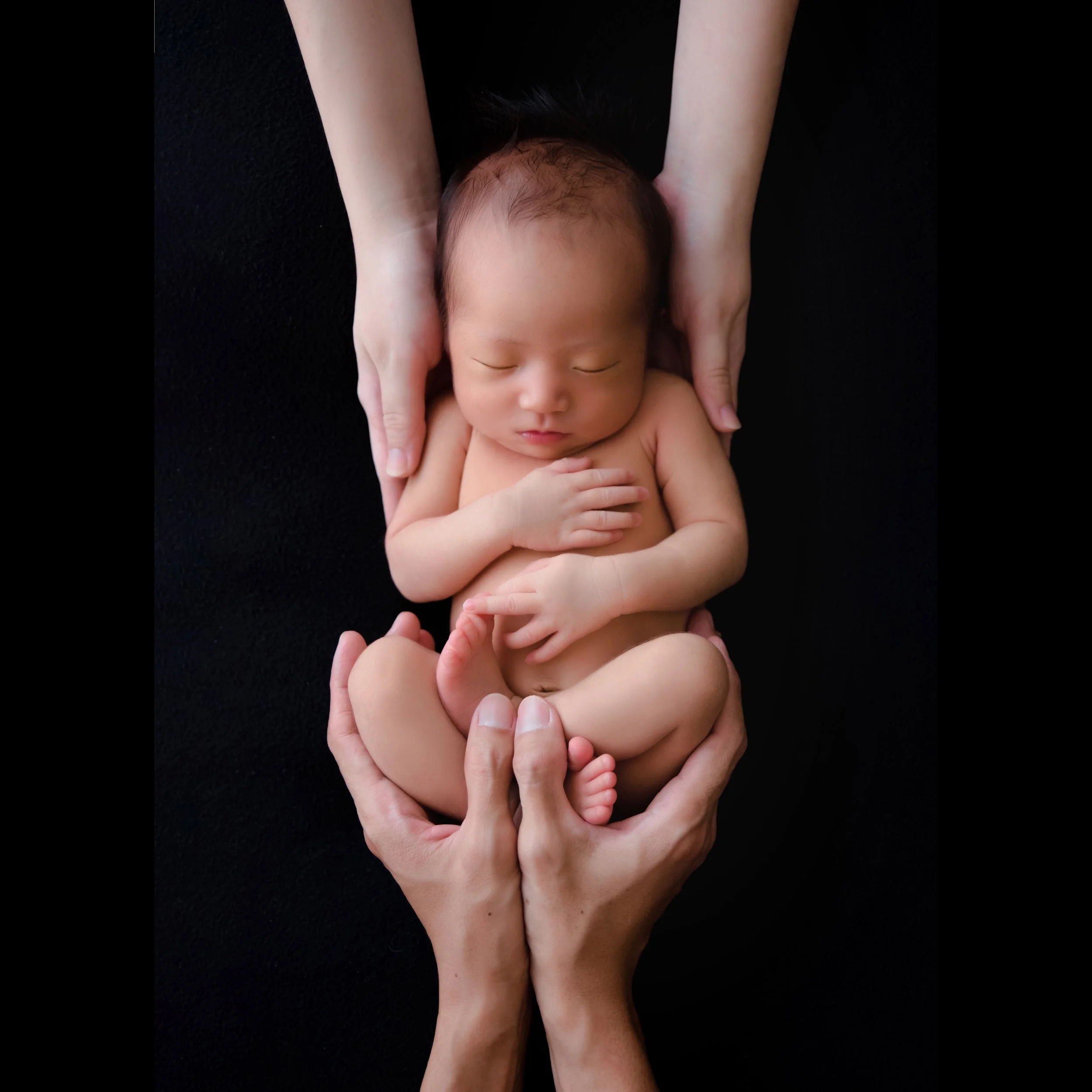 Sleeping newborn curled on one side while supported by several adult hands extending from above and below against a black backdrop