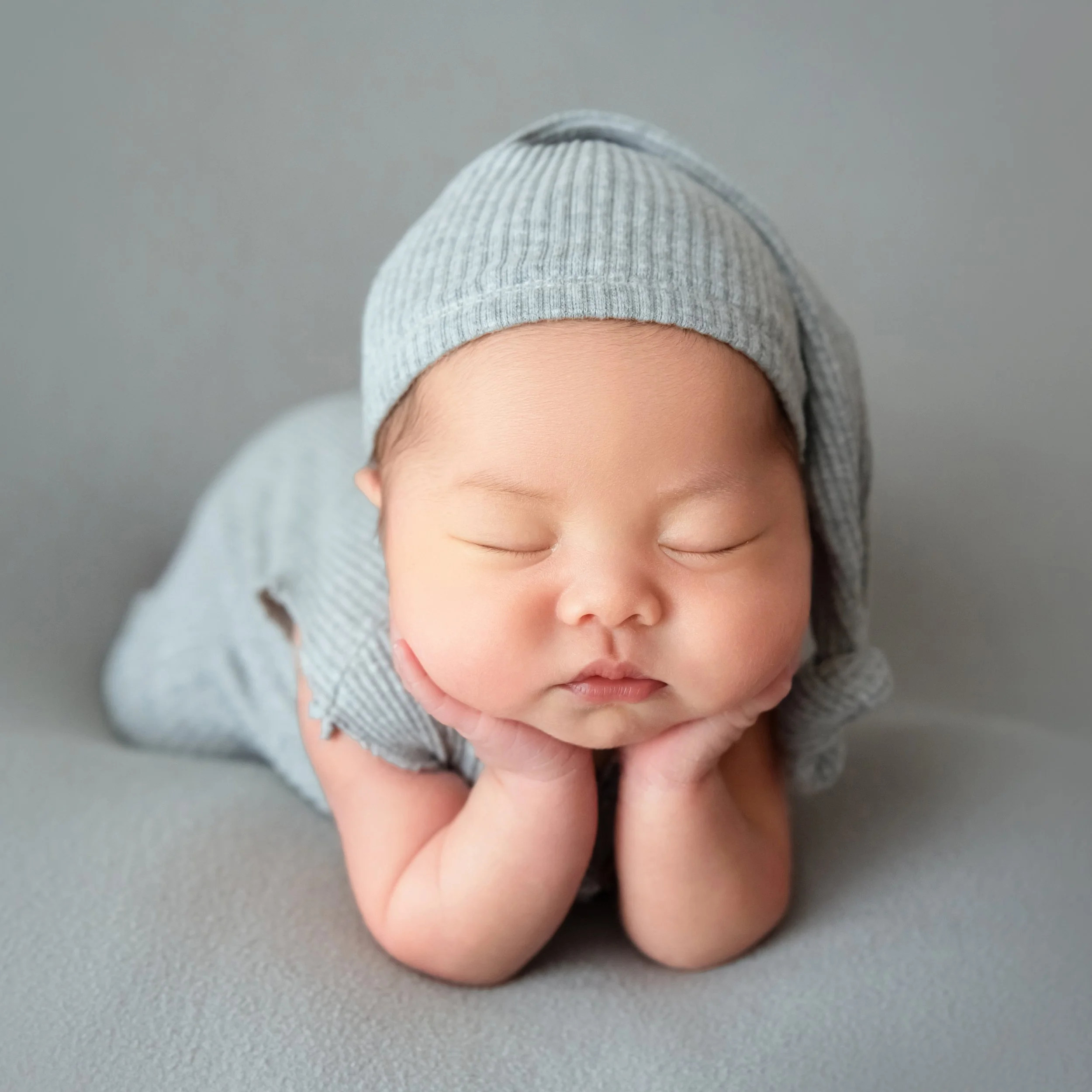 Close-up of a sleeping newborn in a grey knitted hat resting both cheeks on hands on a plain grey backdrop