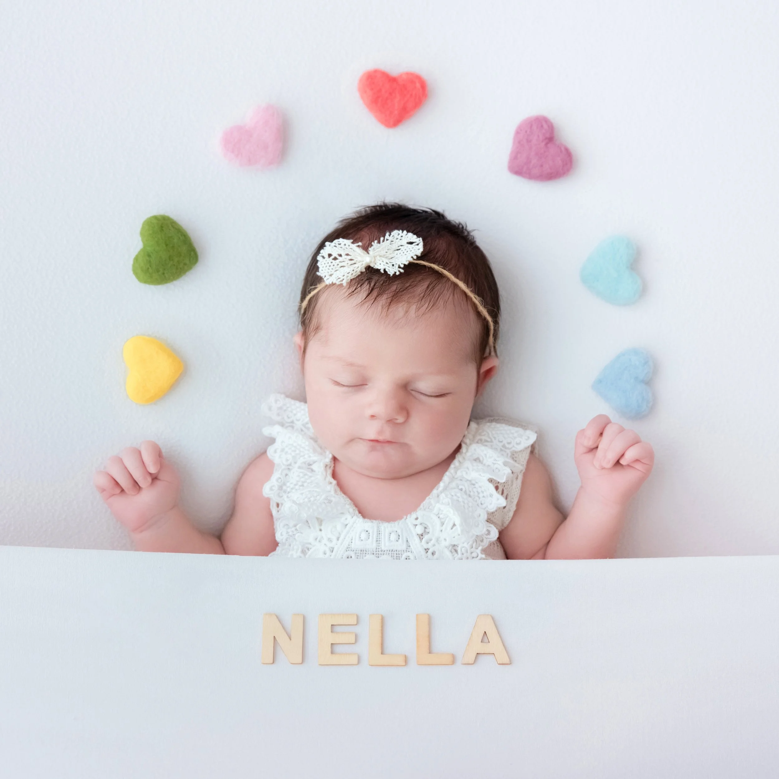 Sleeping newborn in a white lace outfit and bow headband reclining with hands beside the head beneath a semicircle of colourful felt hearts