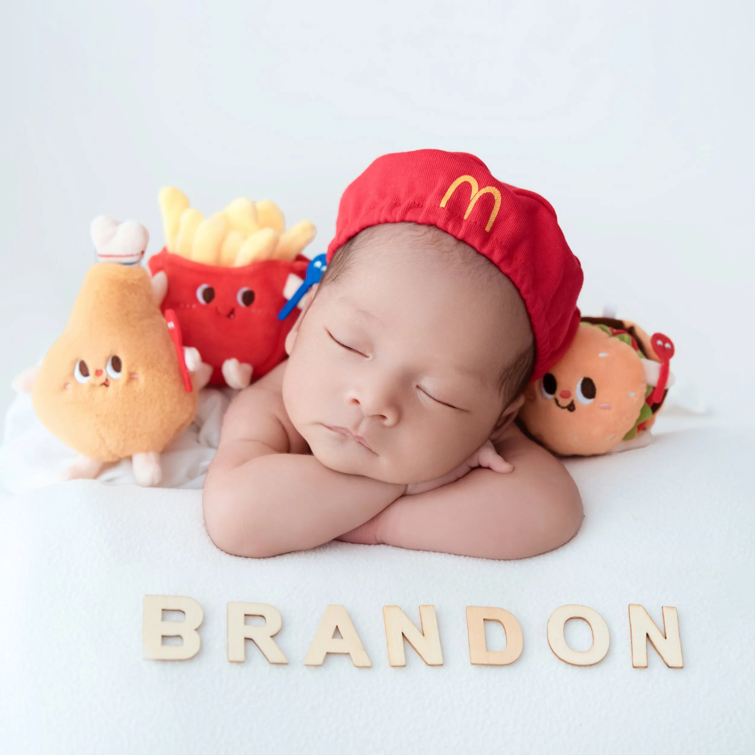 Sleeping newborn in a red McDonald’s cap with chin resting on folded arms, beside fast-food plush toys