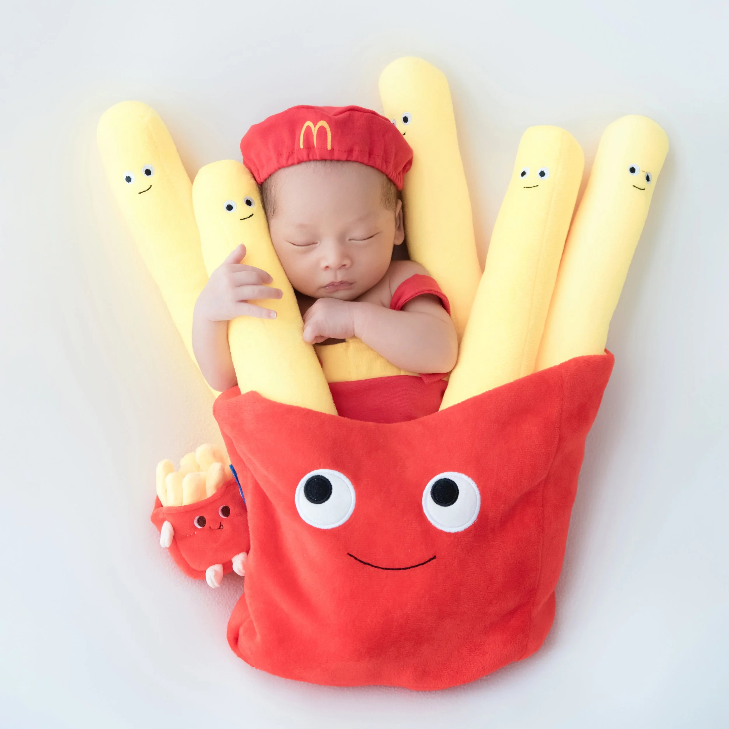 Sleeping newborn in a red McDonald’s cap, nestled in a smiling red fries container with oversized yellow fries