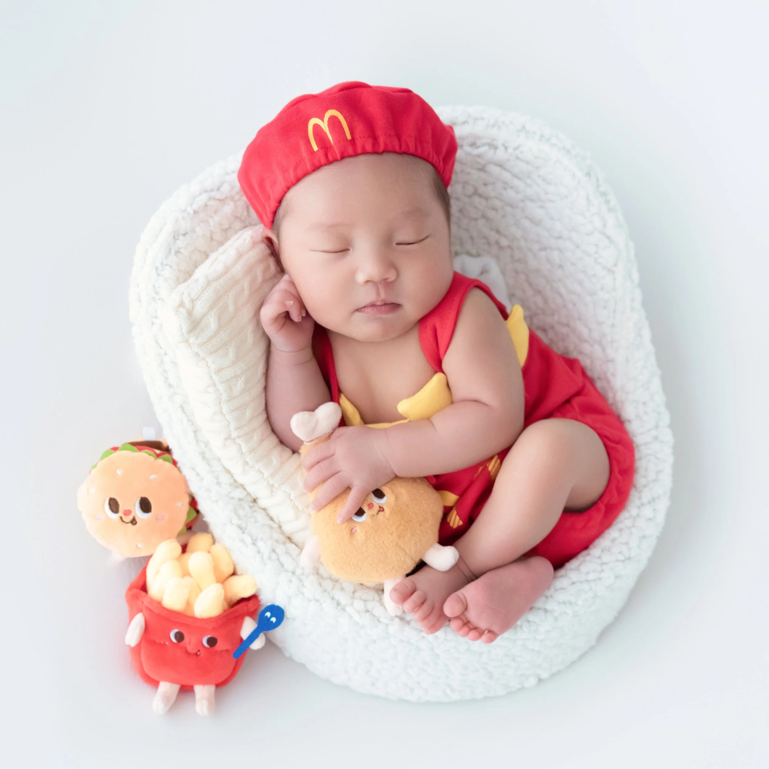 Sleeping newborn in a red McDonald’s cap and fries outfit, curled in a white basket beside fast-food plush toys