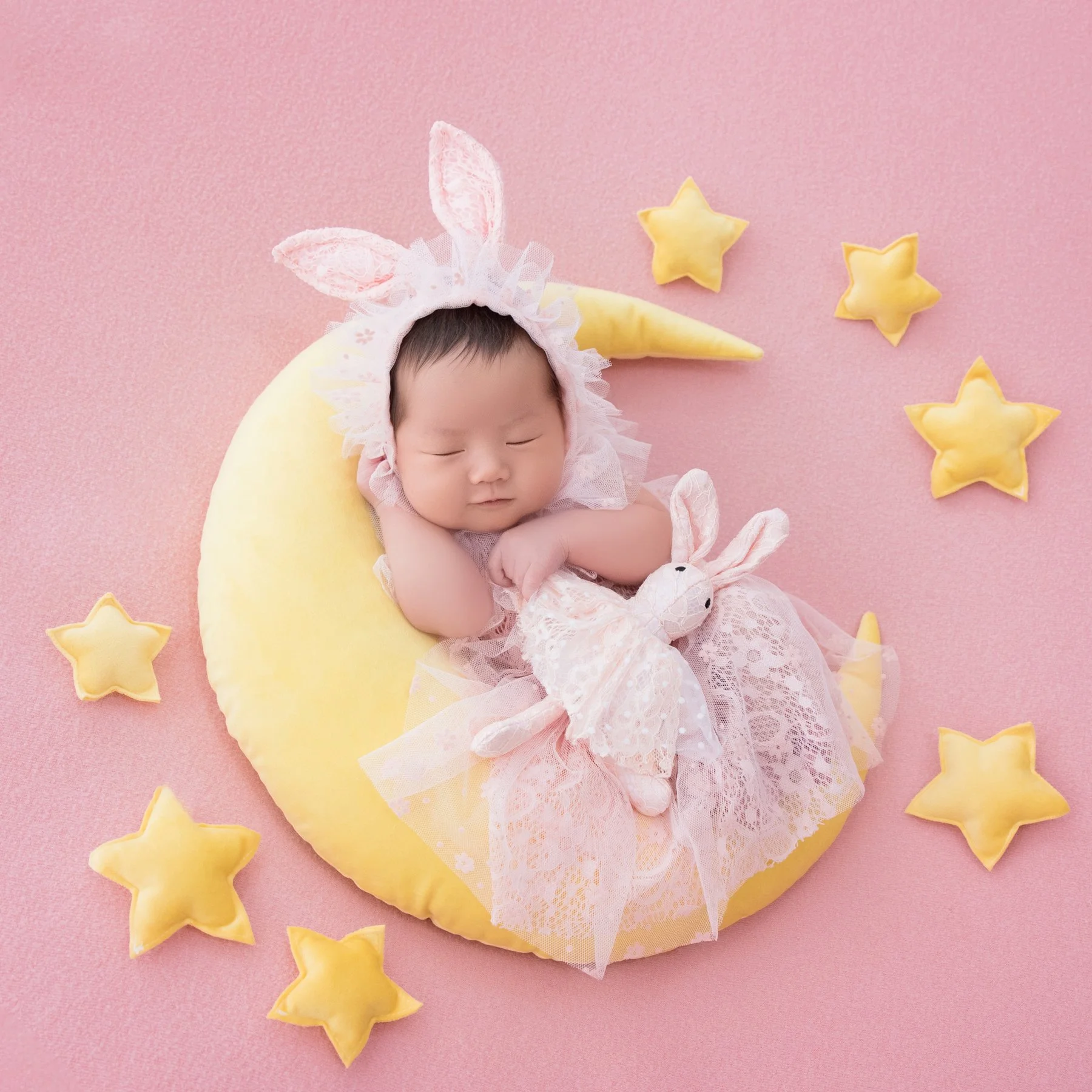 Sleeping newborn in a pink lace dress and bunny-ear headband holding a rabbit plush on a yellow crescent moon