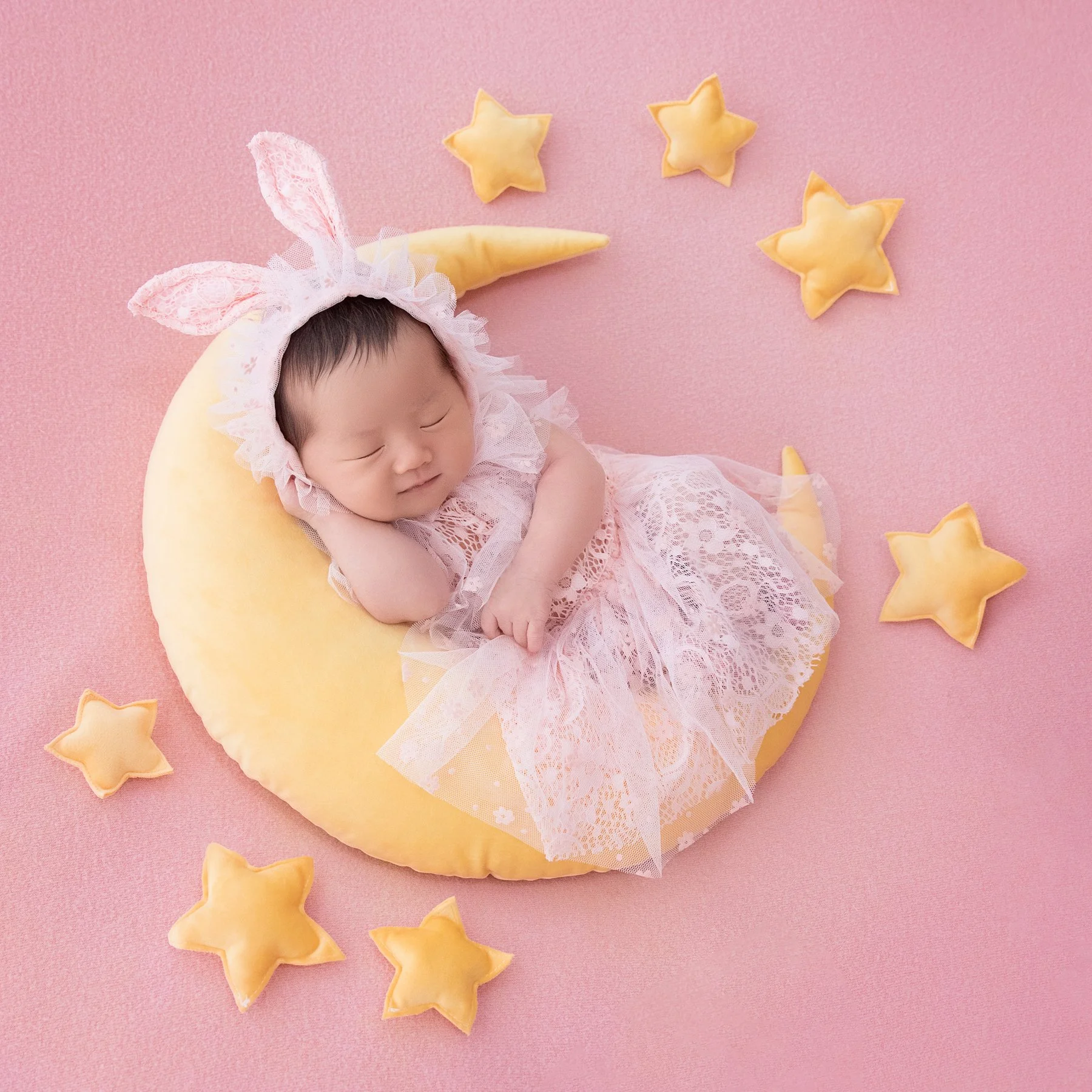 Sleeping newborn in a pink lace dress and bunny-ear headband on a yellow crescent moon surrounded by stars