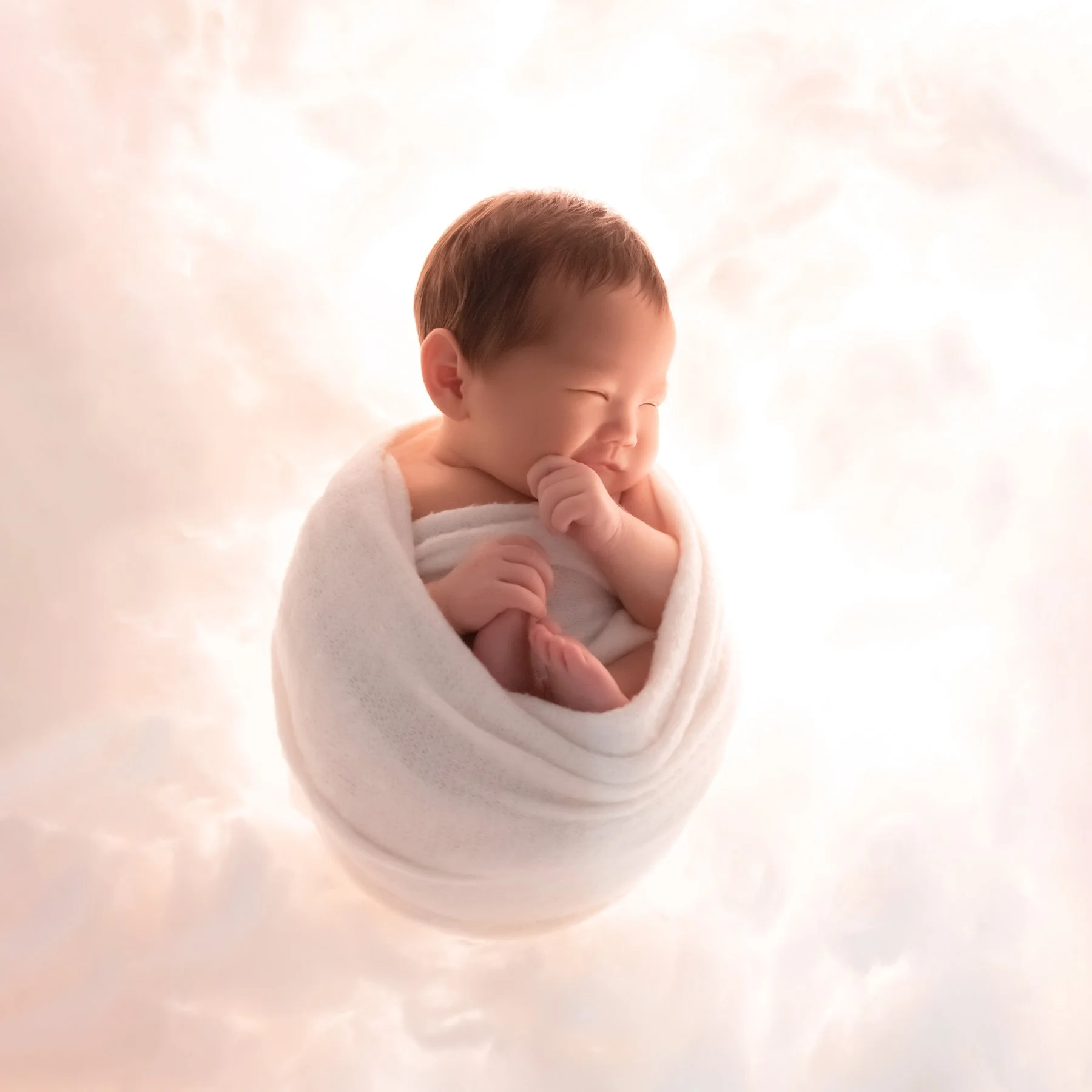 Sleeping newborn curled in a white swaddle with one hand beside the cheek against a softly lit white cloud backdrop