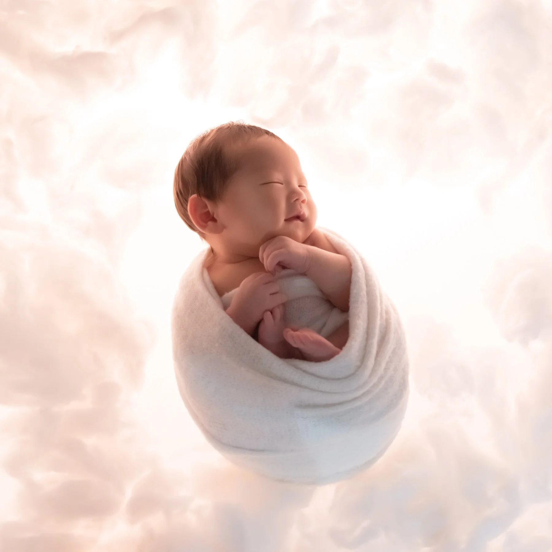 Sleeping newborn curled in a pale blue swaddle with hands near the cheek against a softly lit white cloud backdrop
