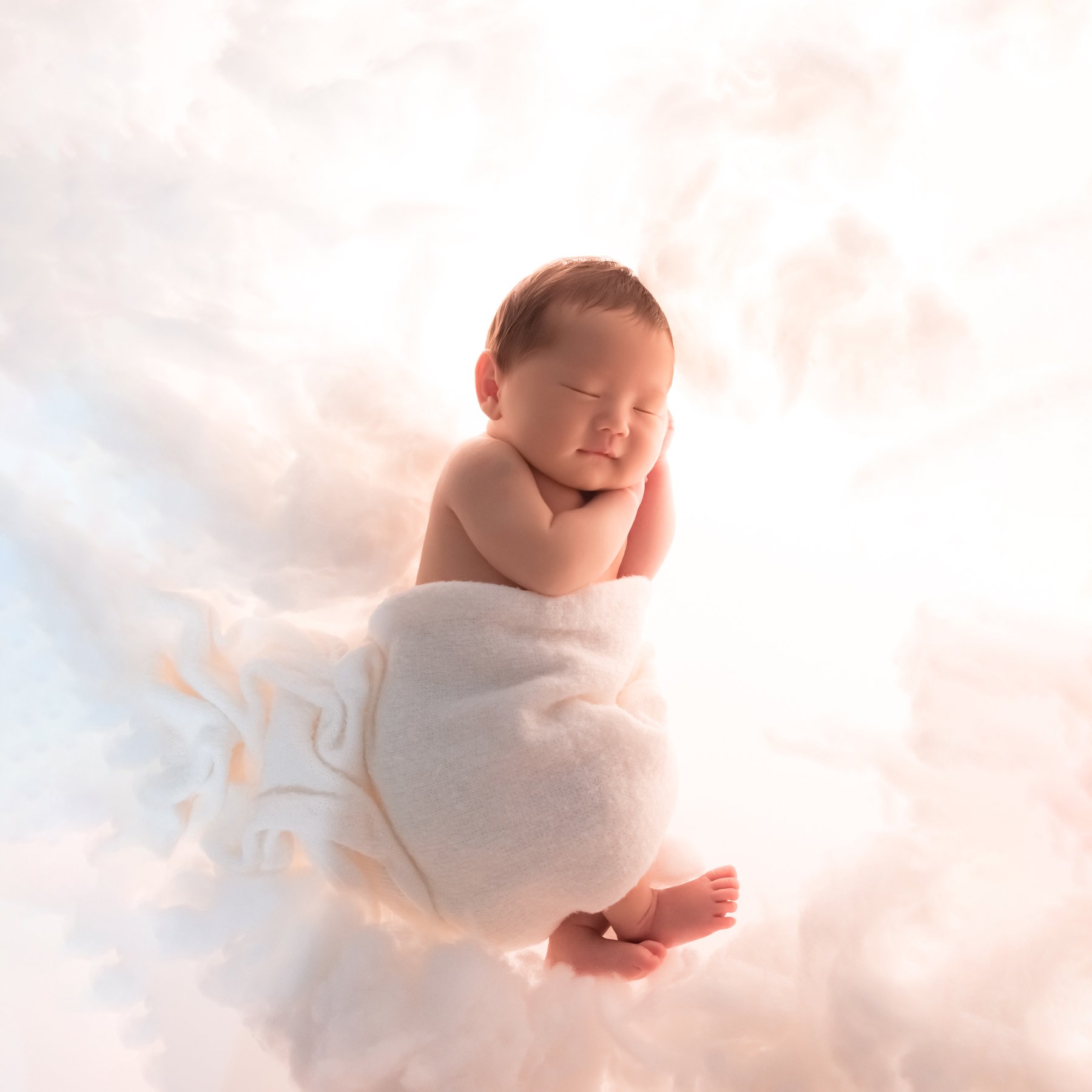 Sleeping newborn wrapped in white fabric with one hand beside the cheek and a tiny foot extending over a softly lit white cloud backdrop