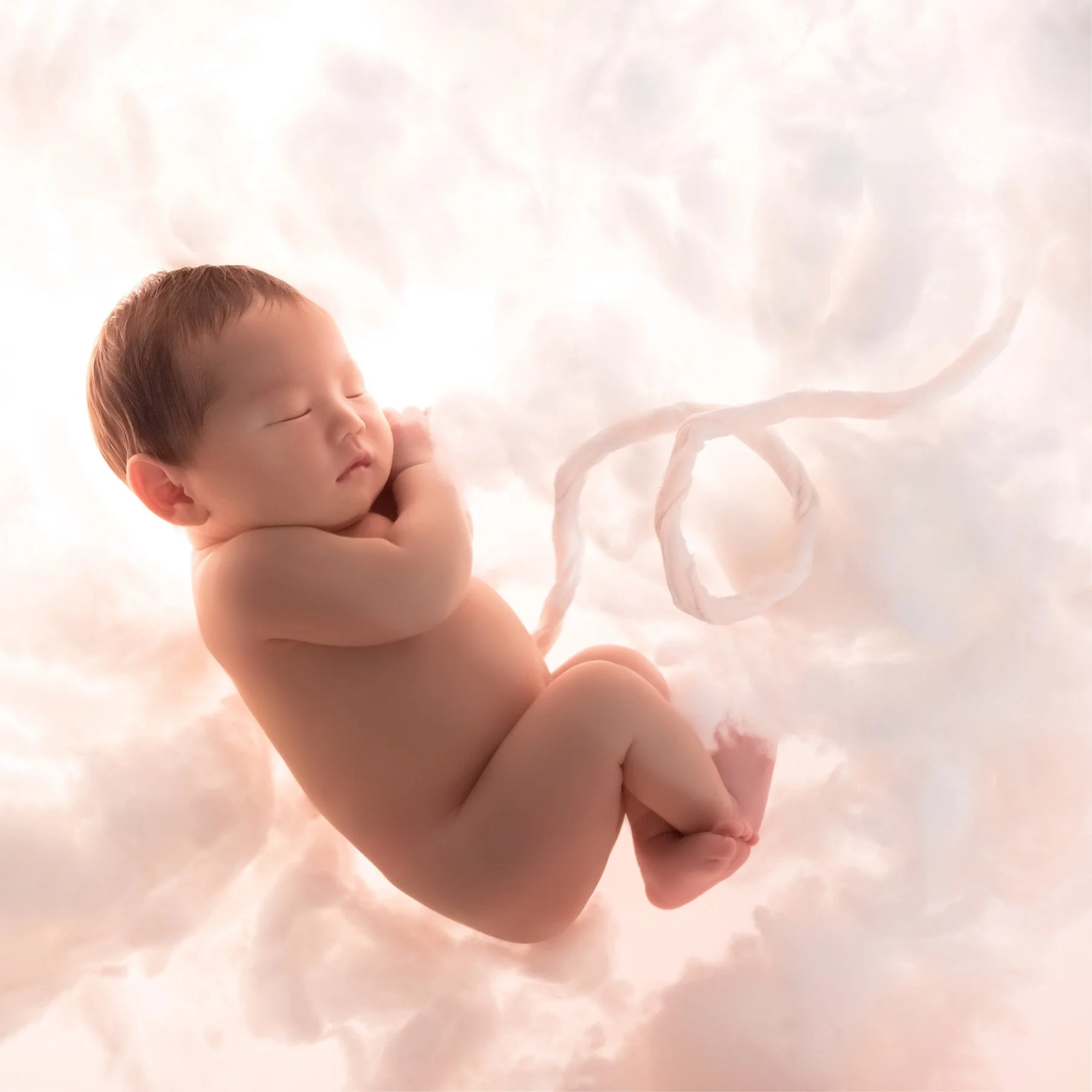 Sleeping newborn curled on one side with hands beneath the cheek and legs tucked up against a softly lit white cloud backdrop