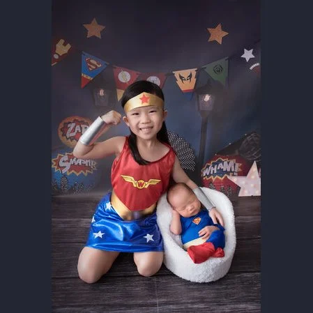 Older sibling in a Wonder Woman outfit kneeling beside a newborn in a Superman costume on a comic-themed backdrop