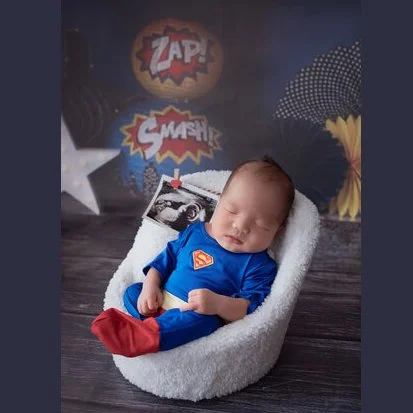 Sleeping newborn in a blue Superman outfit and red socks resting in a white basket against a comic-style backdrop