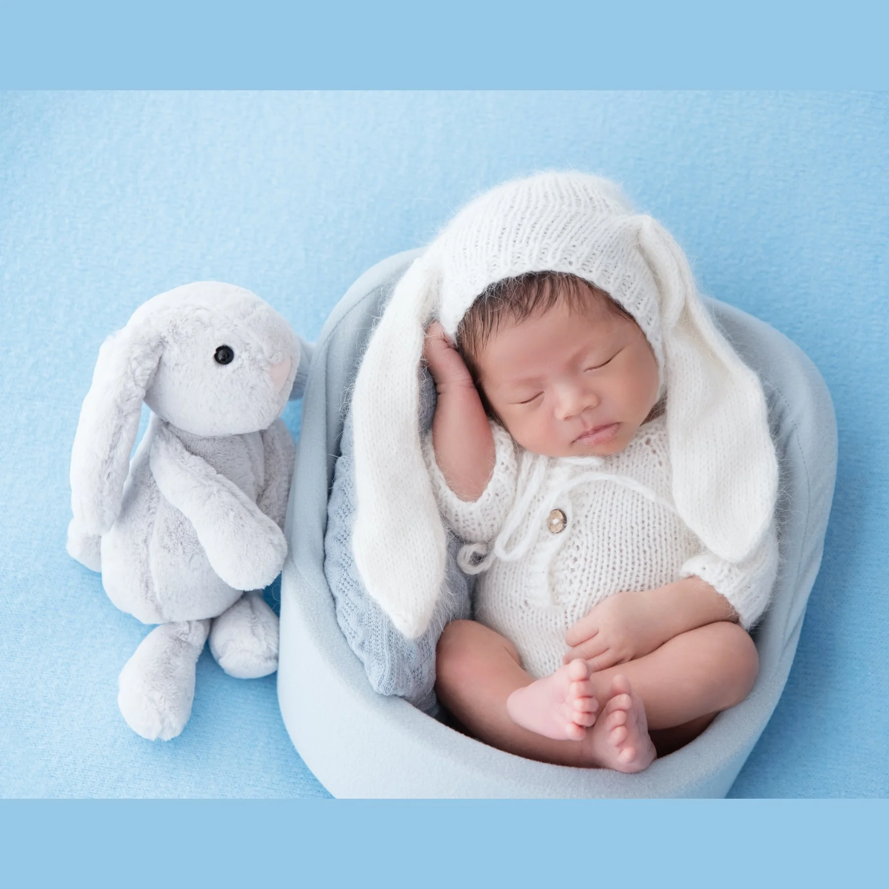 Sleeping newborn in a white floppy-eared bunny hat and knitted romper seated in a blue basket beside a rabbit plush