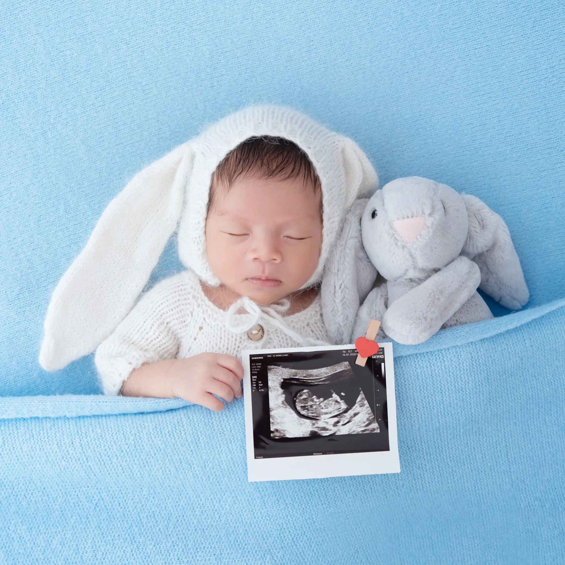 Sleeping newborn in a white floppy-eared bunny hat beside a grey rabbit plush against a blue backdrop