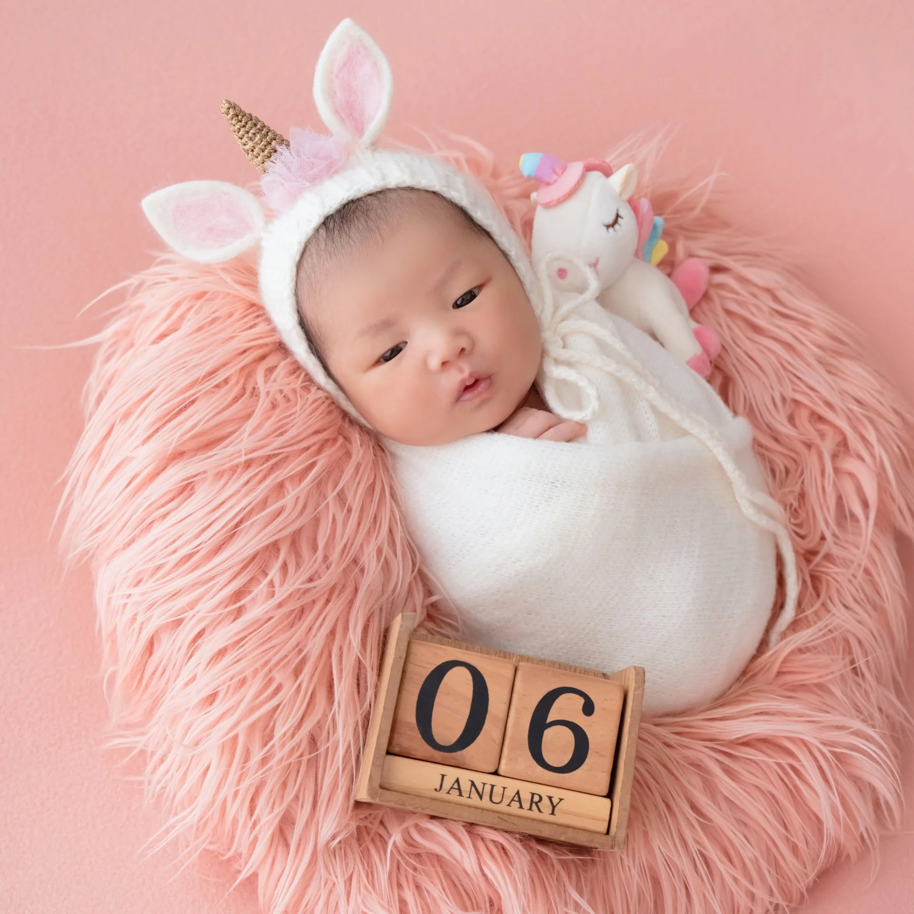 Awake newborn in a white wrap and unicorn hat lying on pink fur with a small unicorn toy beside the head