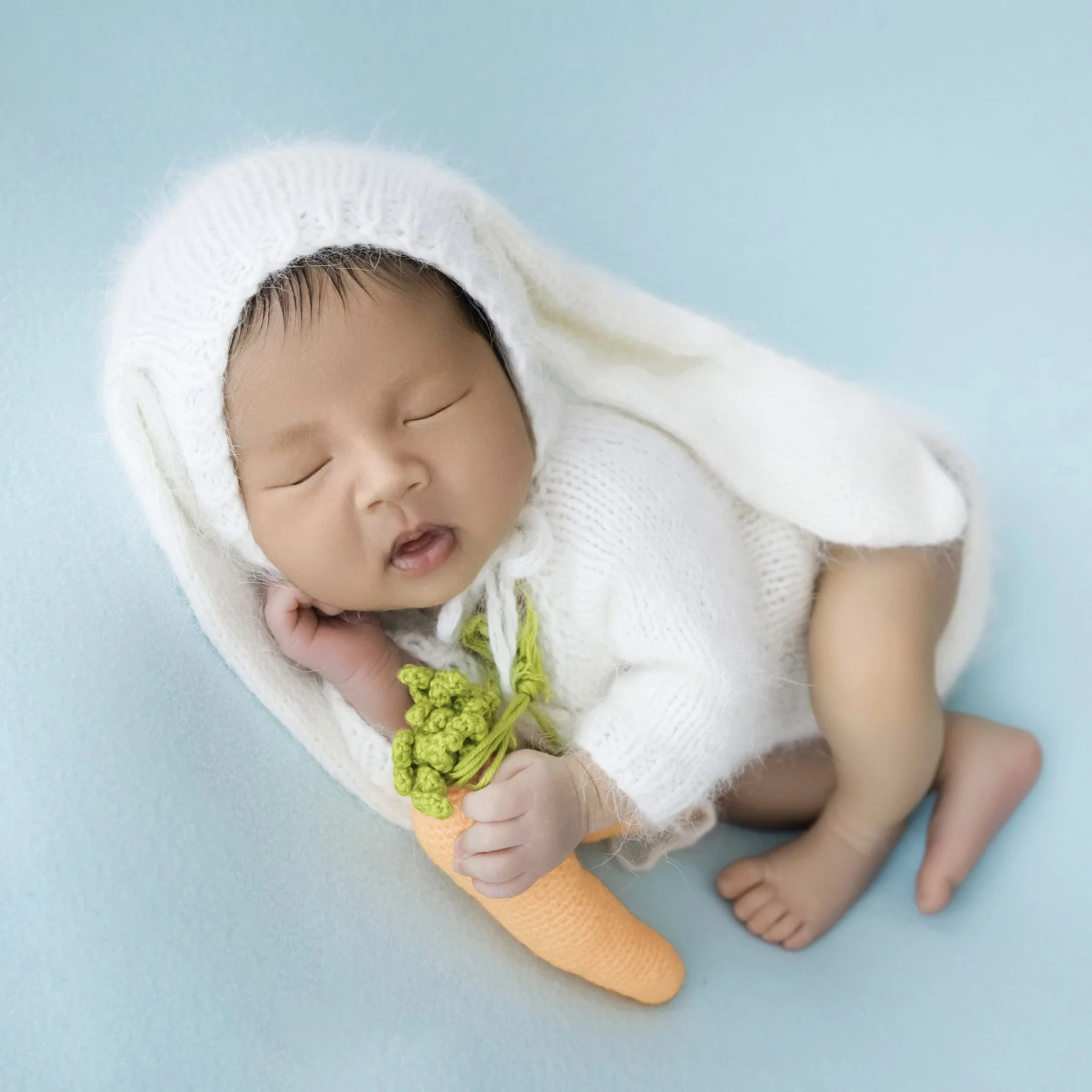 Smiling newborn in a white floppy-eared bunny hat holding a plush carrot while curled on a mint backdrop