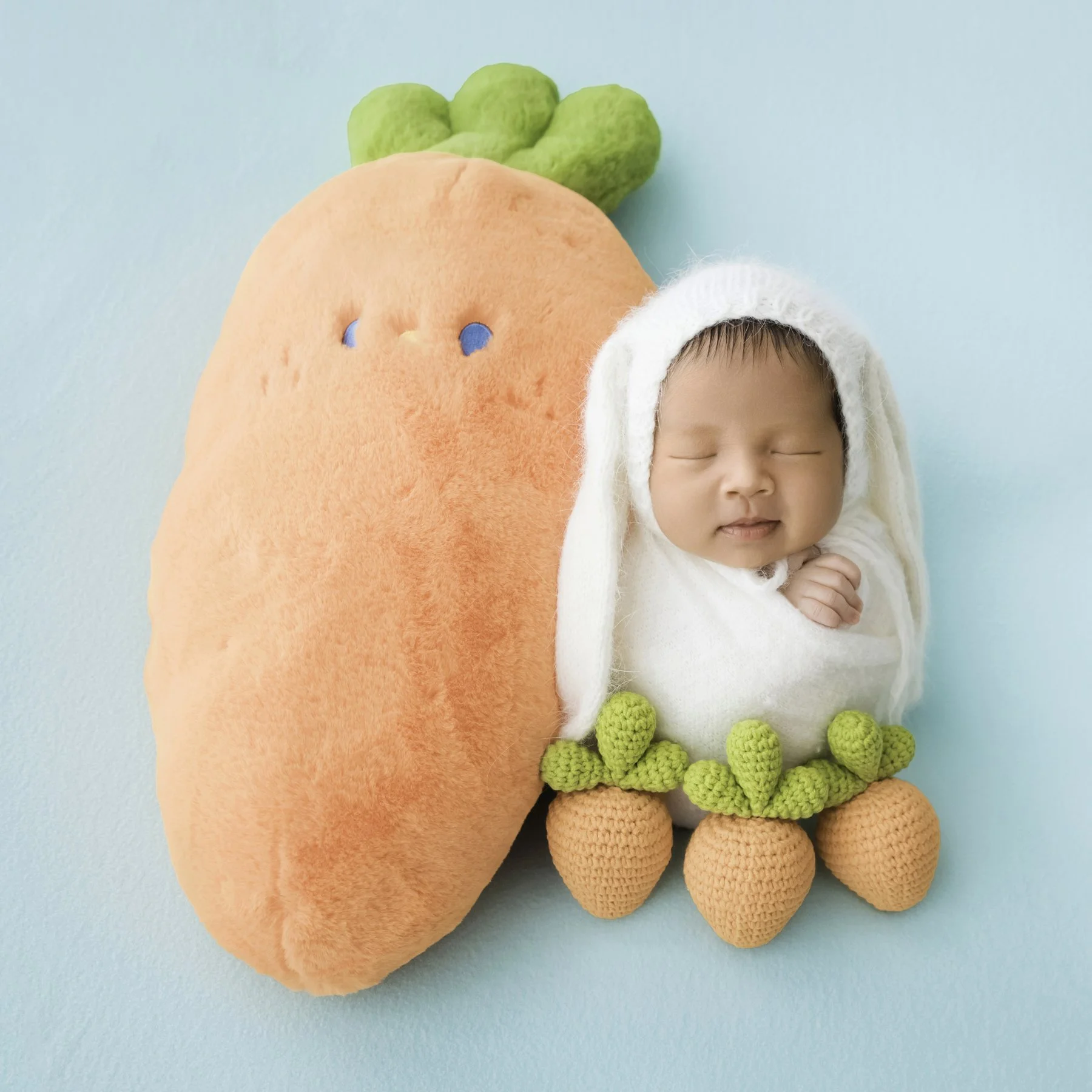Sleeping newborn in a white bunny hat beside a large orange carrot plush and three small carrots on a mint backdrop
