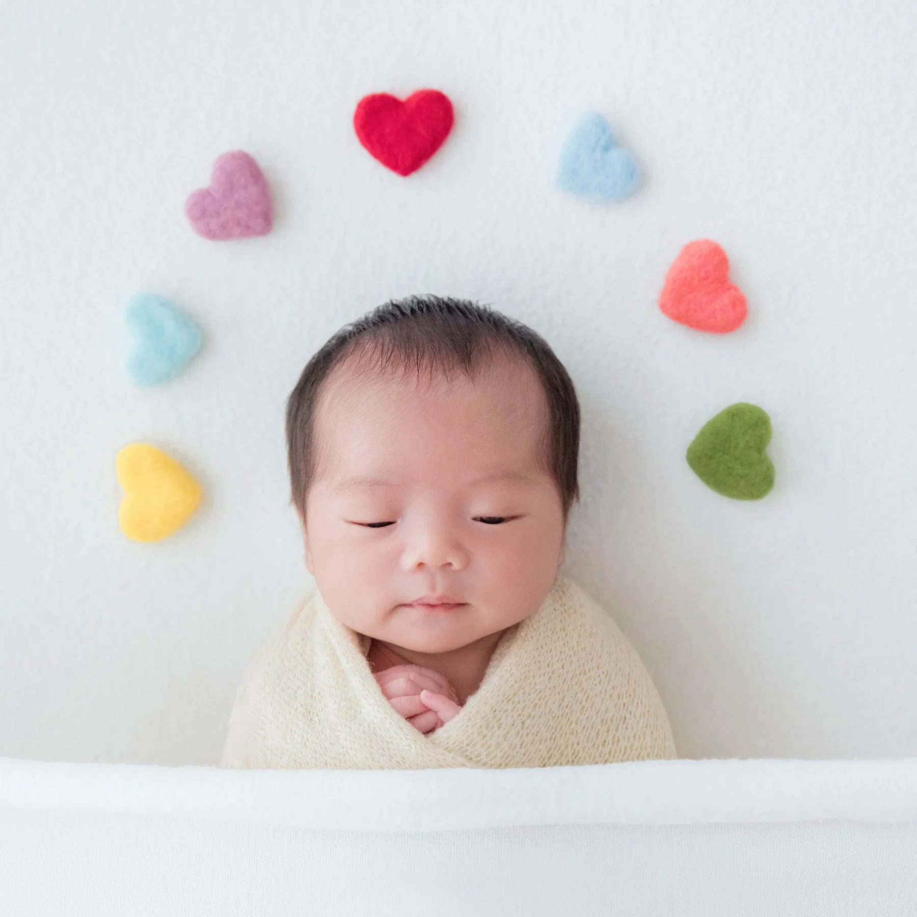 Sleeping newborn in a cream swaddle reclining beneath a semicircle of colourful felt hearts on a white backdrop