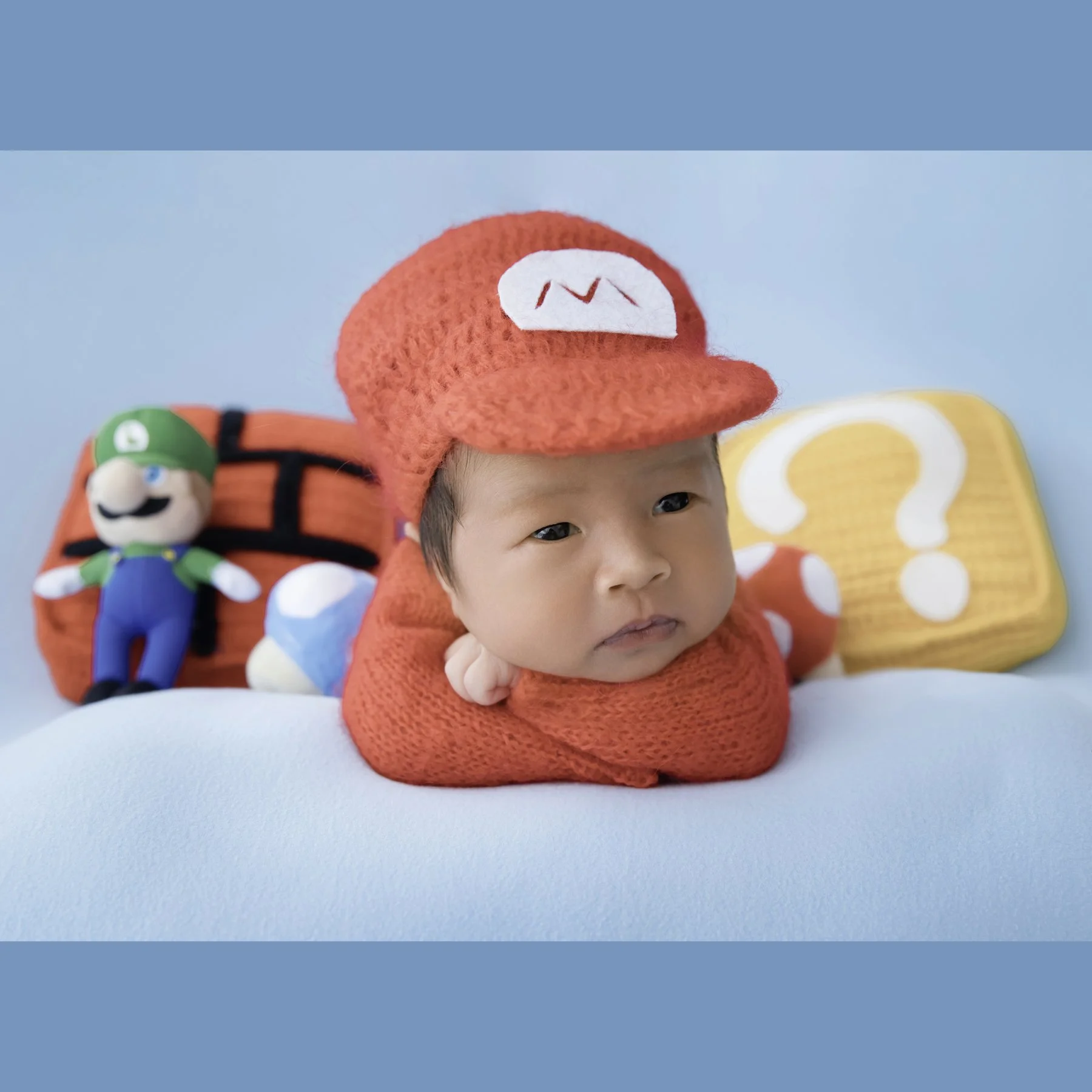 Awake newborn in a red Mario hat resting the chin on folded arms with Luigi and game-themed props behind