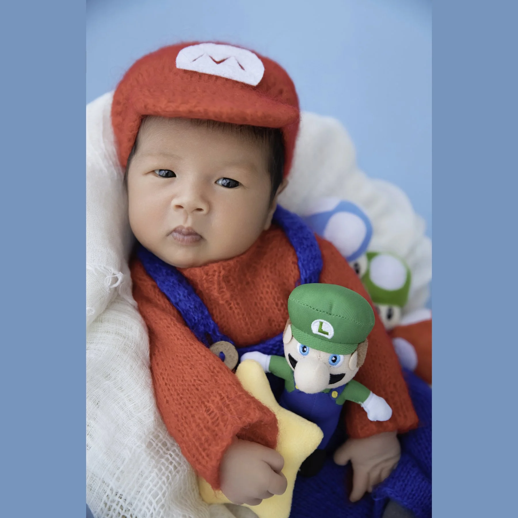 Awake newborn wearing a red Mario hat and knitted overalls, holding a Luigi toy in a white-lined basket