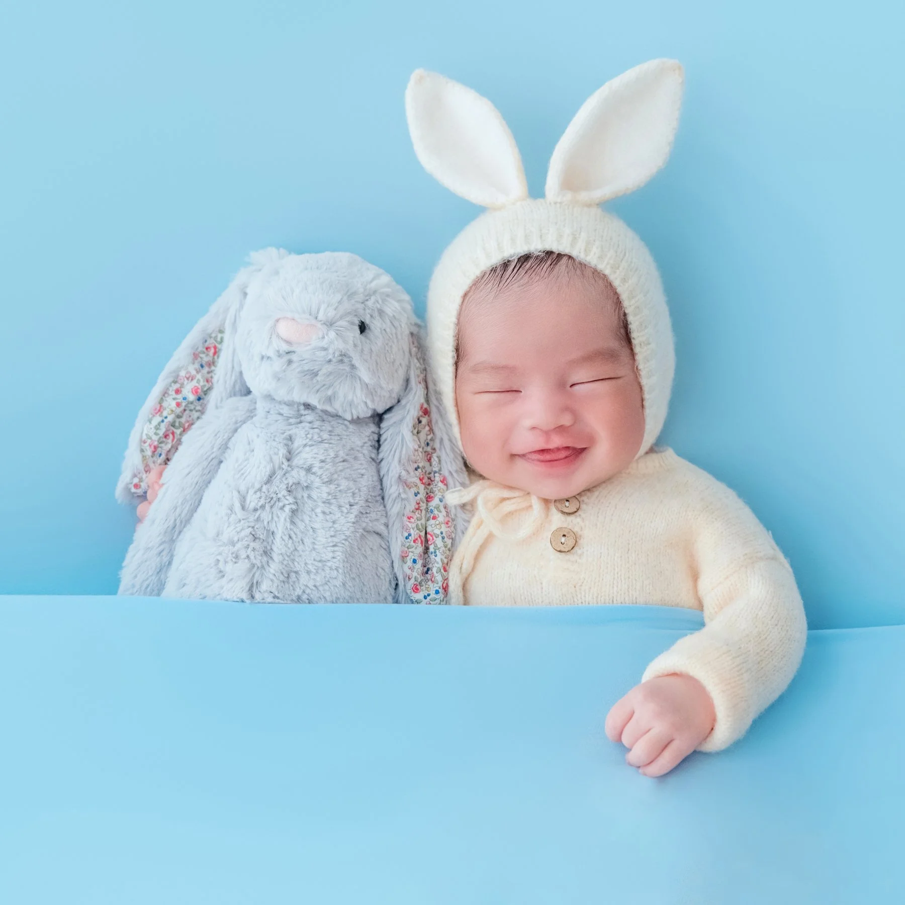 Smiling newborn in a cream knitted bunny hat beside a grey rabbit plush against a blue backdrop