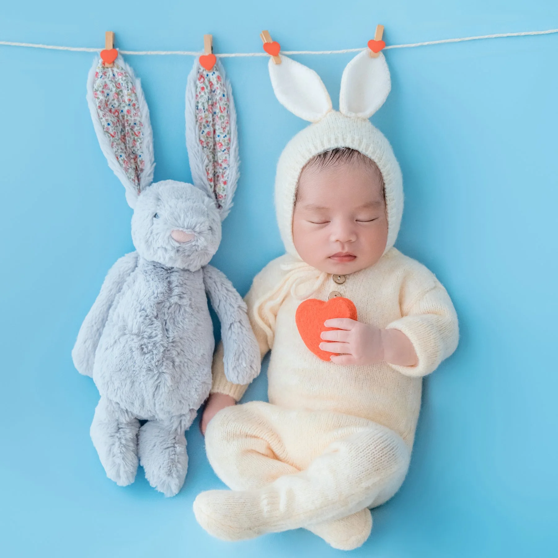 Sleeping newborn in a cream knitted bunny outfit holding a carrot prop beside a grey rabbit plush against blue