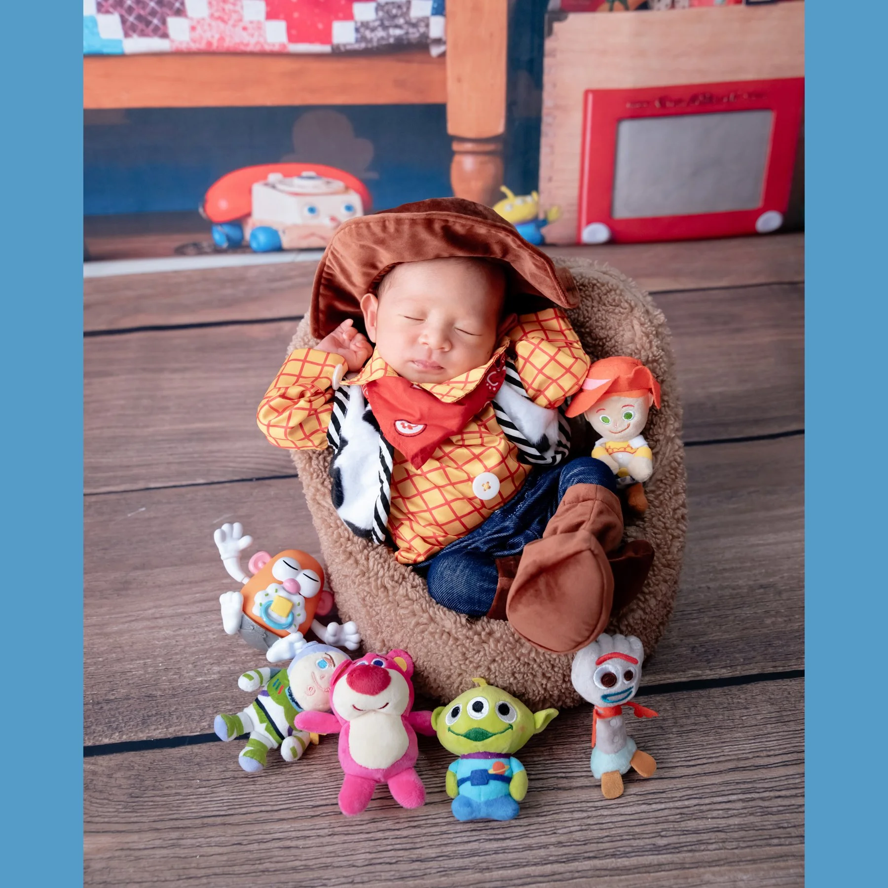 Sleeping newborn in a Woody costume with both hands beside the head in a basket surrounded by colourful Toy Story toys
