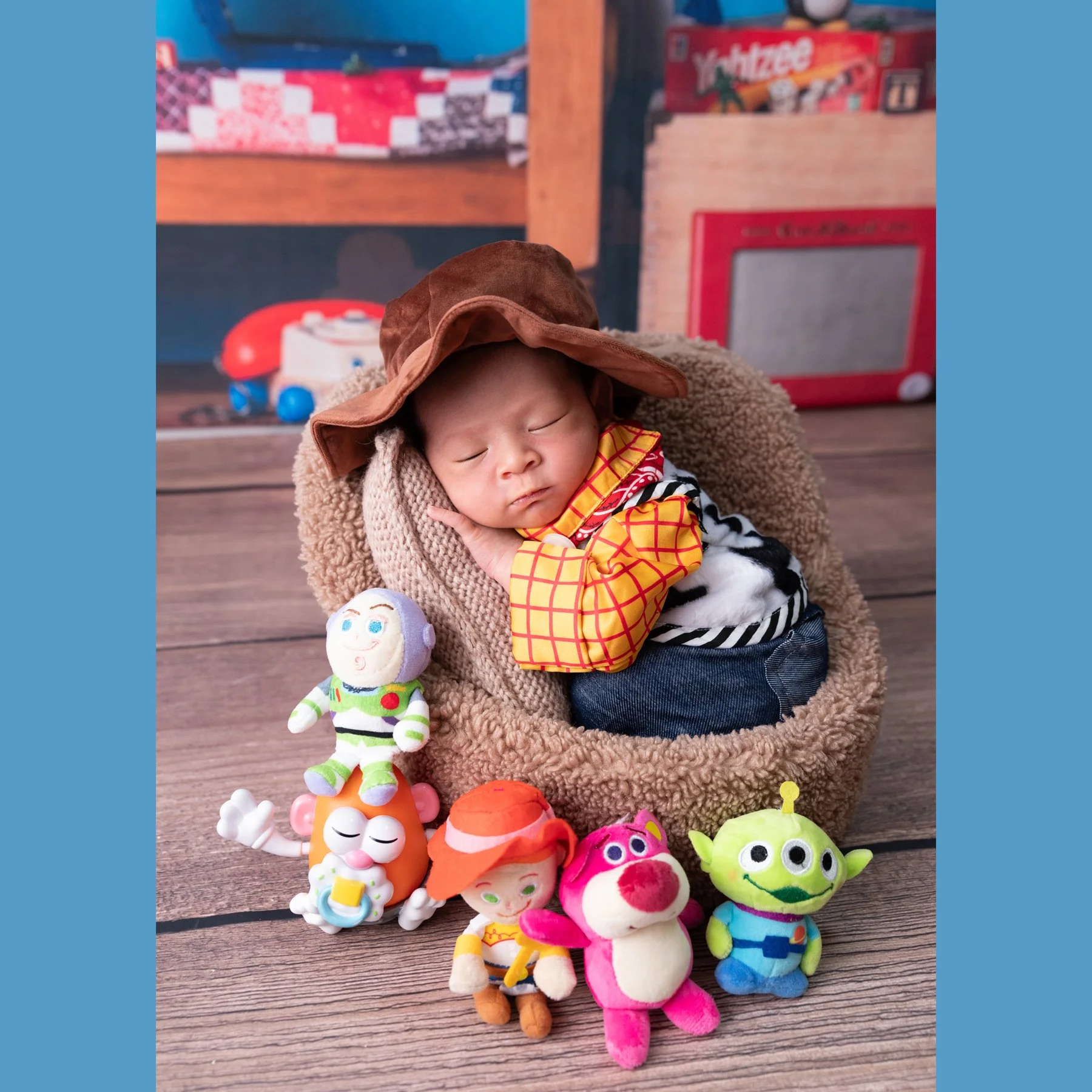 Sleeping newborn in a Woody cowboy outfit curled in a basket with one hand beside the face and Toy Story toys below