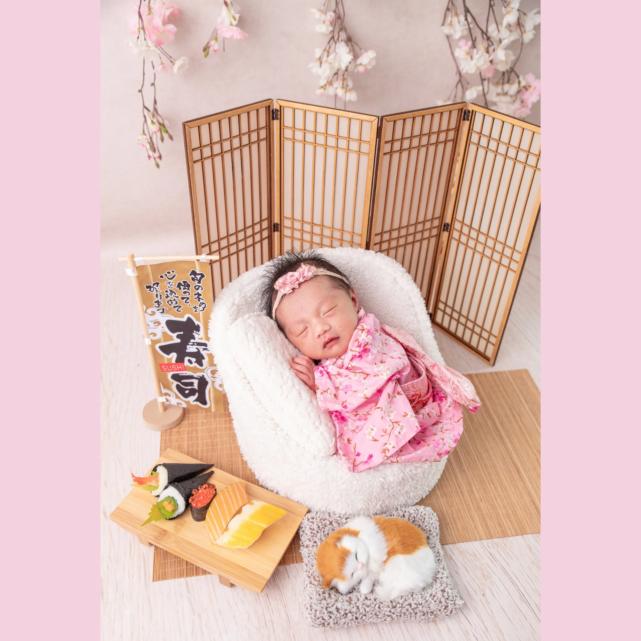 Sleeping newborn in a pink floral kimono and matching headband reclining in a white basket beside toy sushi and a wooden Japanese screen
