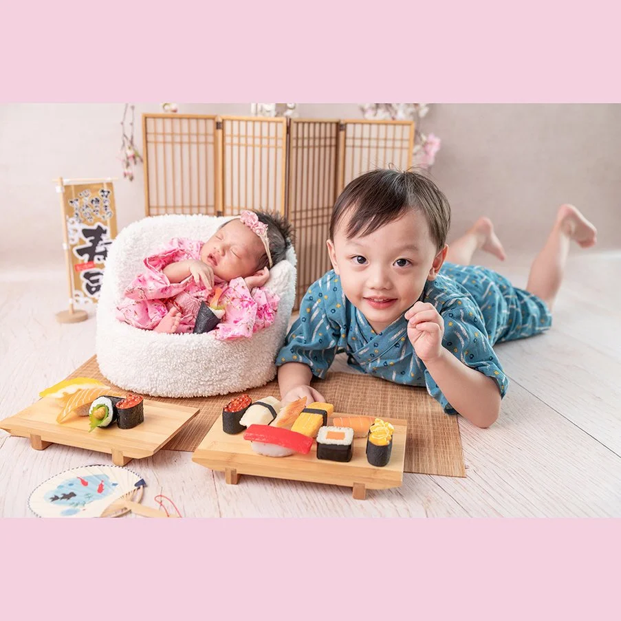 Smiling older sibling in a blue patterned outfit lying beside a sleeping newborn in a pink kimono with toy sushi and a wooden Japanese screen