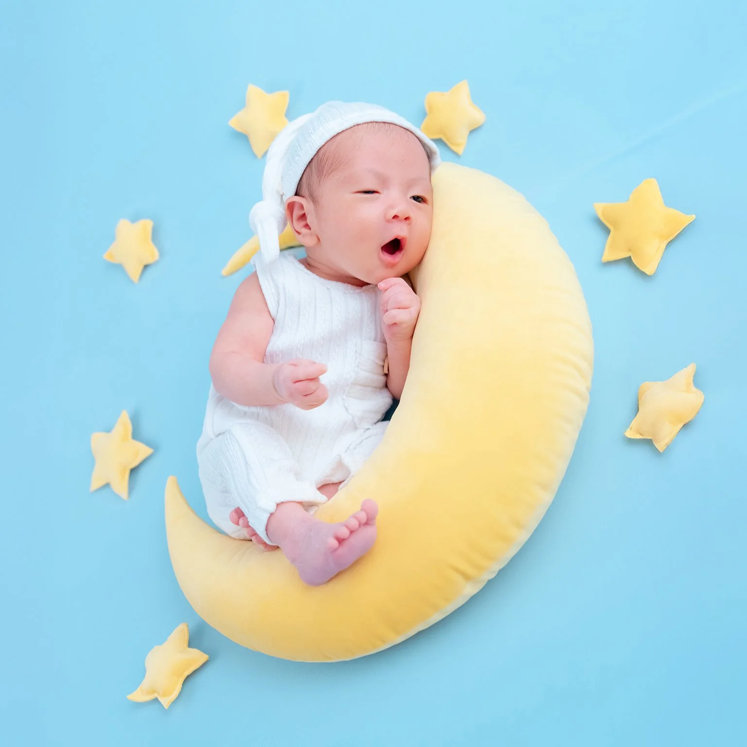 Yawning newborn in white clothing resting on a yellow crescent moon with stars