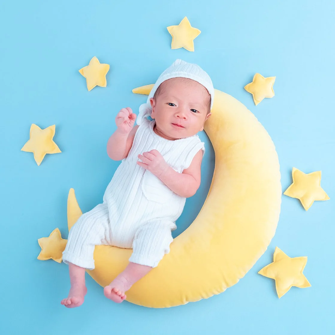 Awake newborn in white clothing lying on a yellow crescent moon surrounded by stars