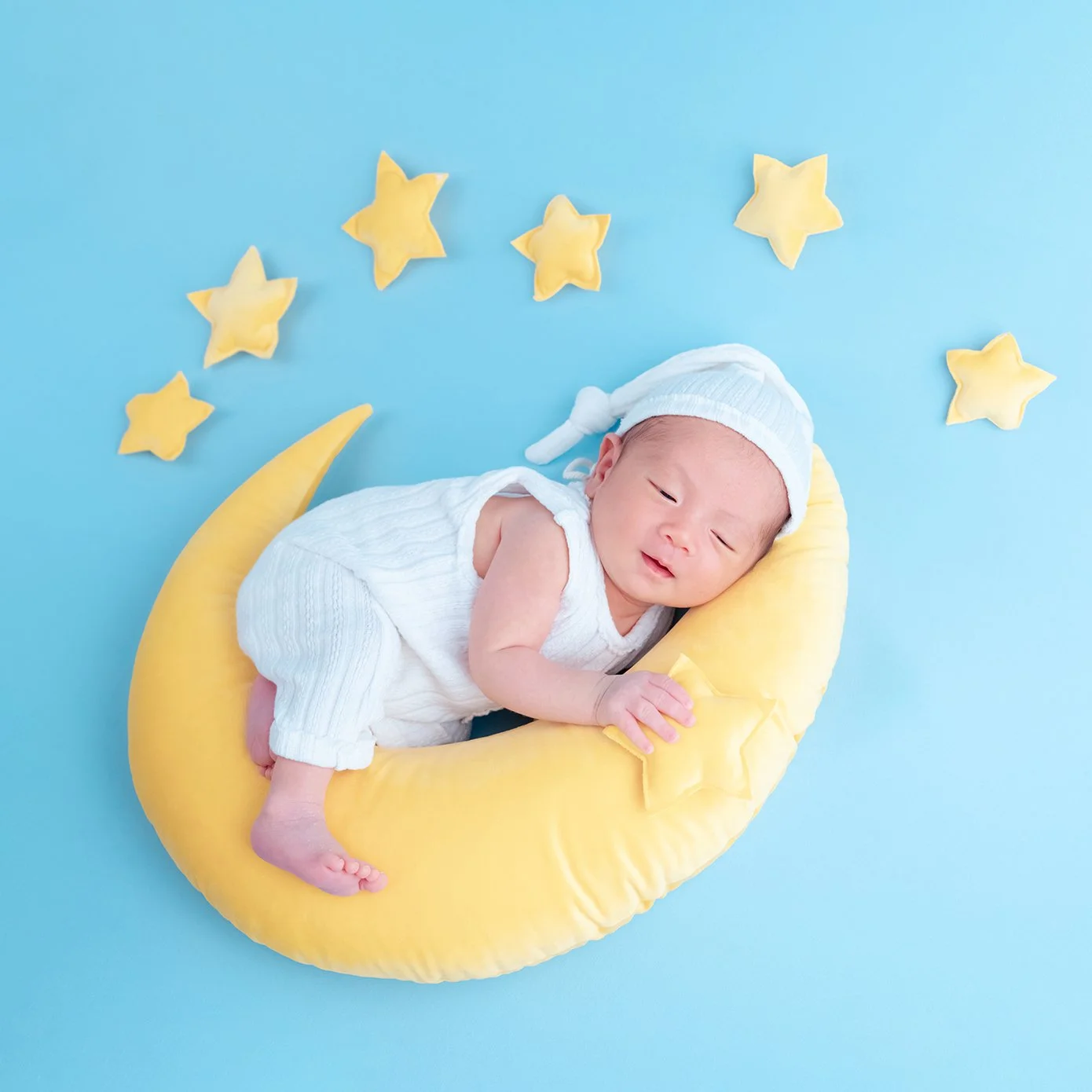 Newborn in a white bonnet curled on a yellow crescent moon against a blue starry backdrop