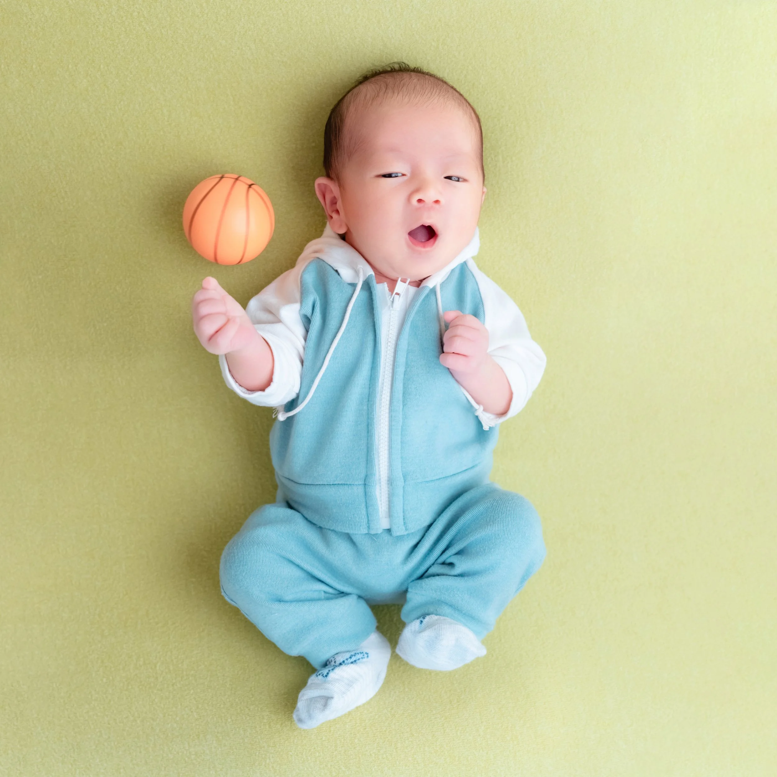 Yawning newborn in a turquoise tracksuit lying on a green blanket with a miniature basketball beside the head
