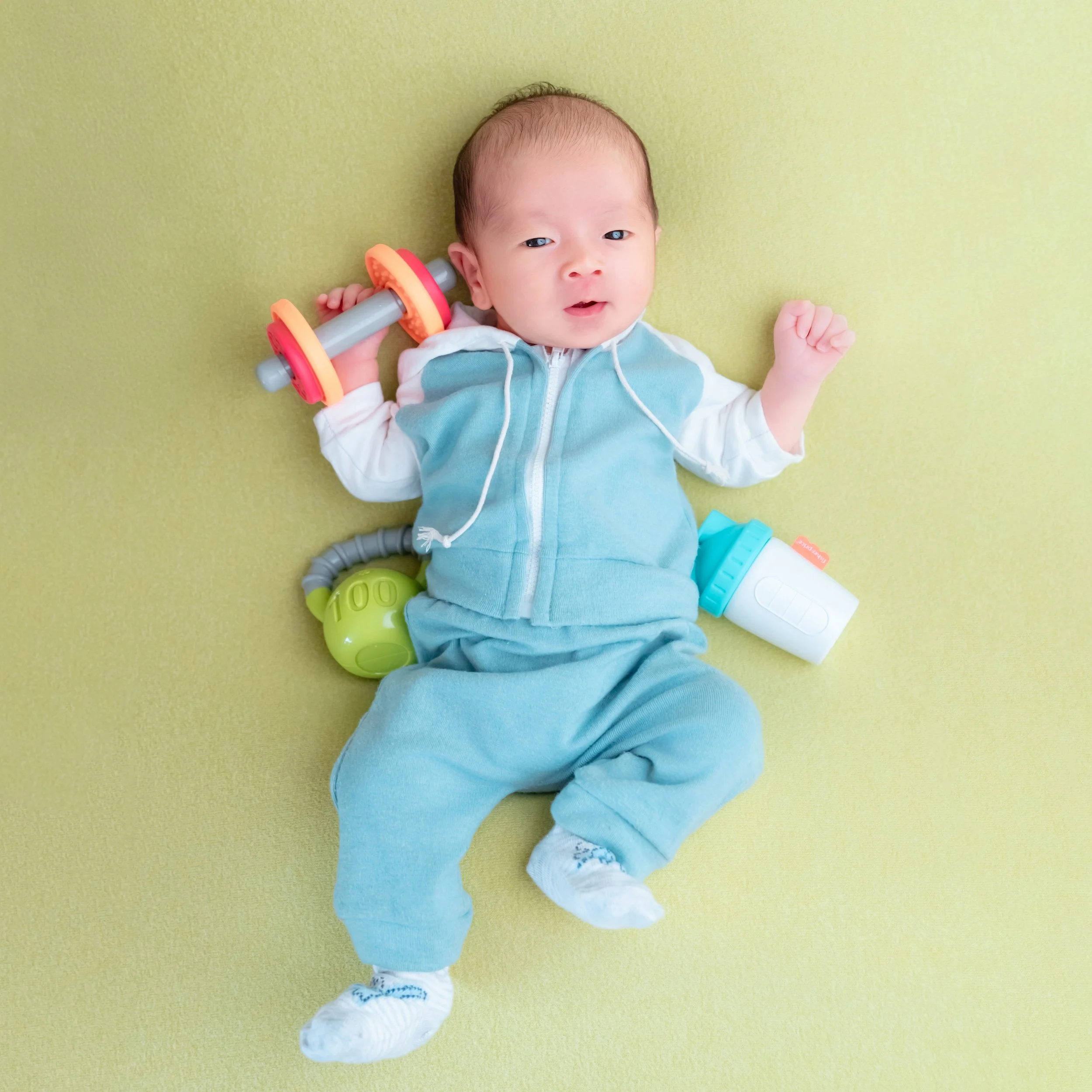 Awake newborn in a turquoise tracksuit lying on a green blanket beside a toy dumbbell, tennis ball and drink bottle