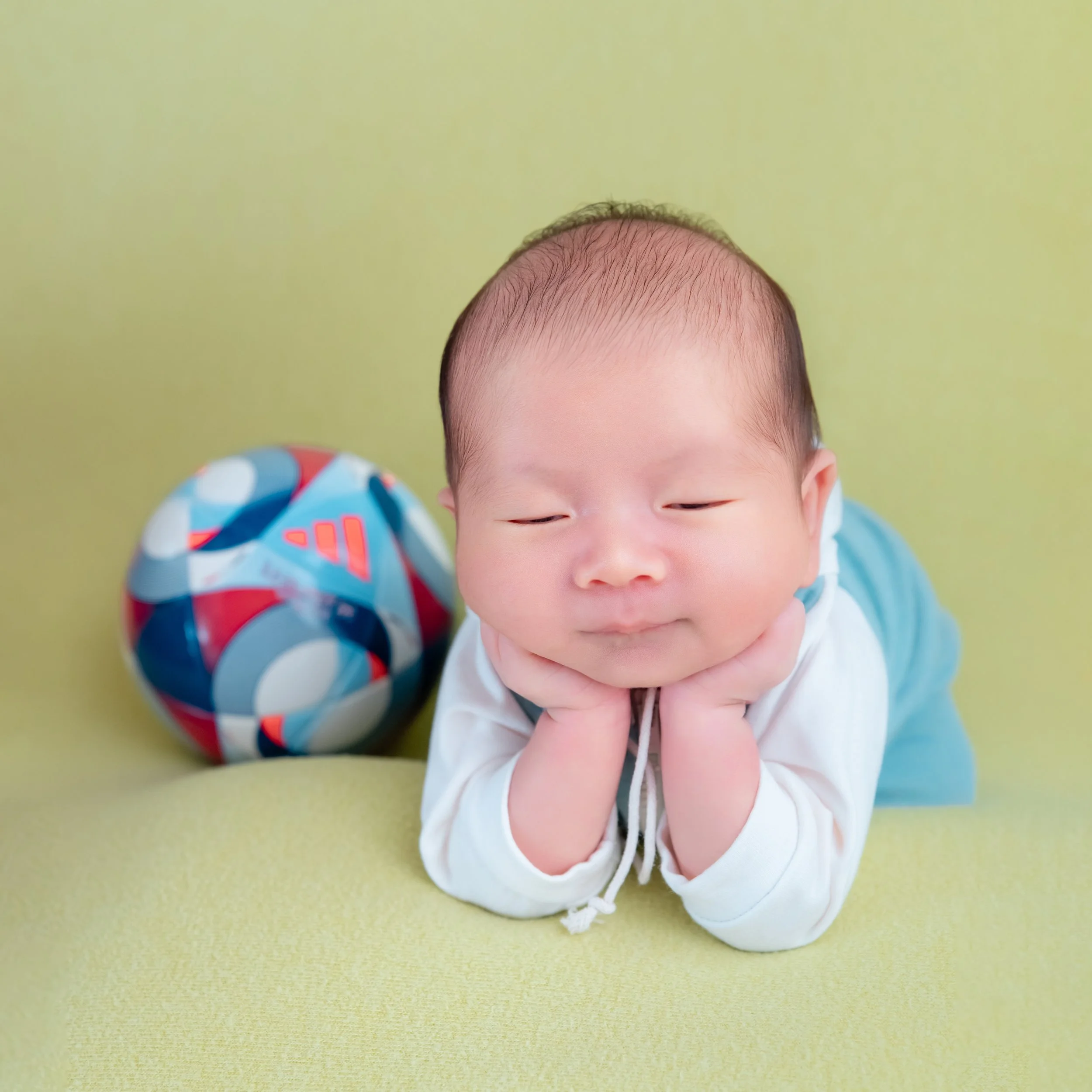 Close-up of a sleeping newborn in a turquoise tracksuit resting both cheeks on hands beside a colourful football on a green blanket