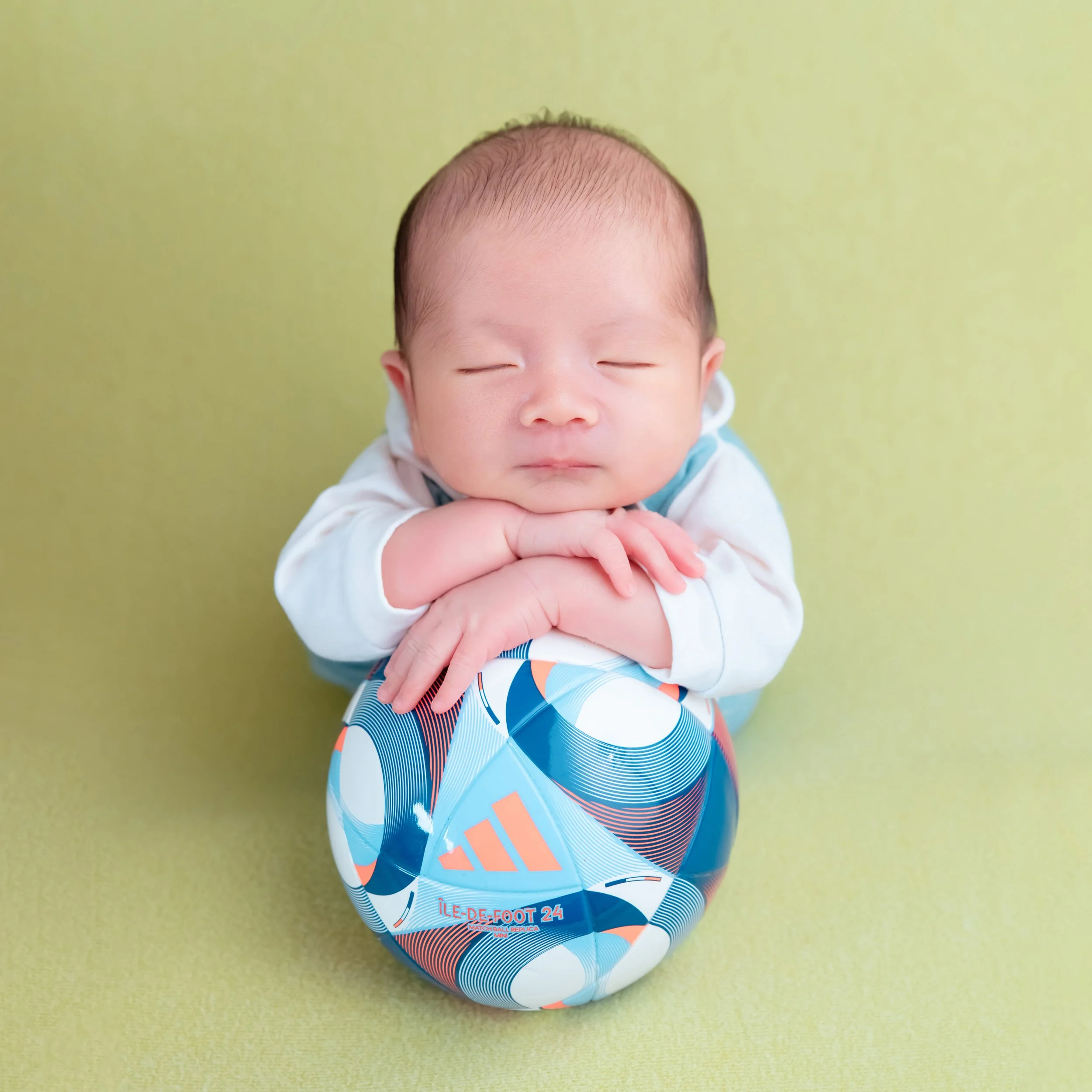 Sleeping newborn in a turquoise tracksuit resting the chin on folded hands above a colourful football on a green blanket