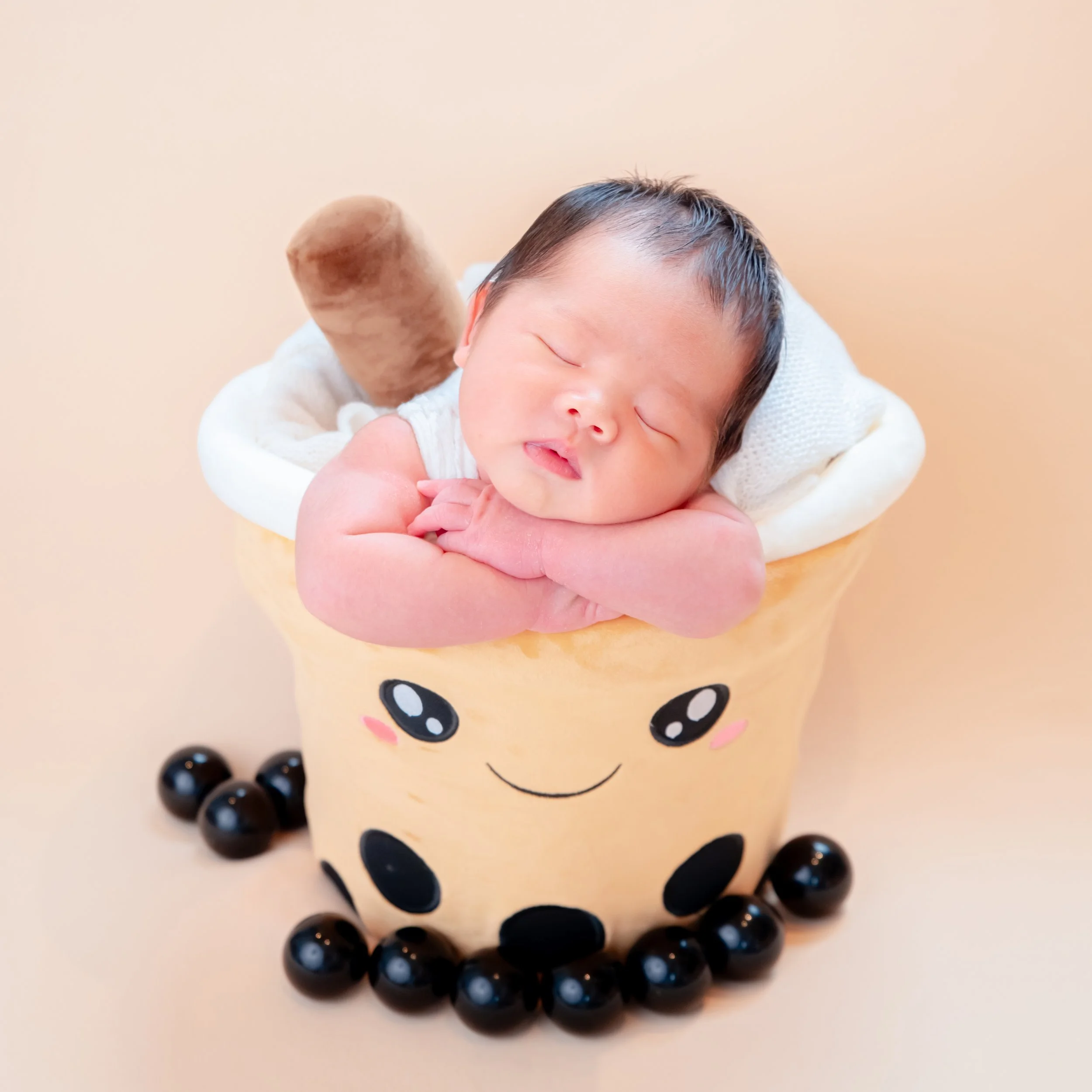 Sleeping newborn resting folded arms on a smiling bubble-tea cup prop, surrounded by black pearls on a peach backdrop