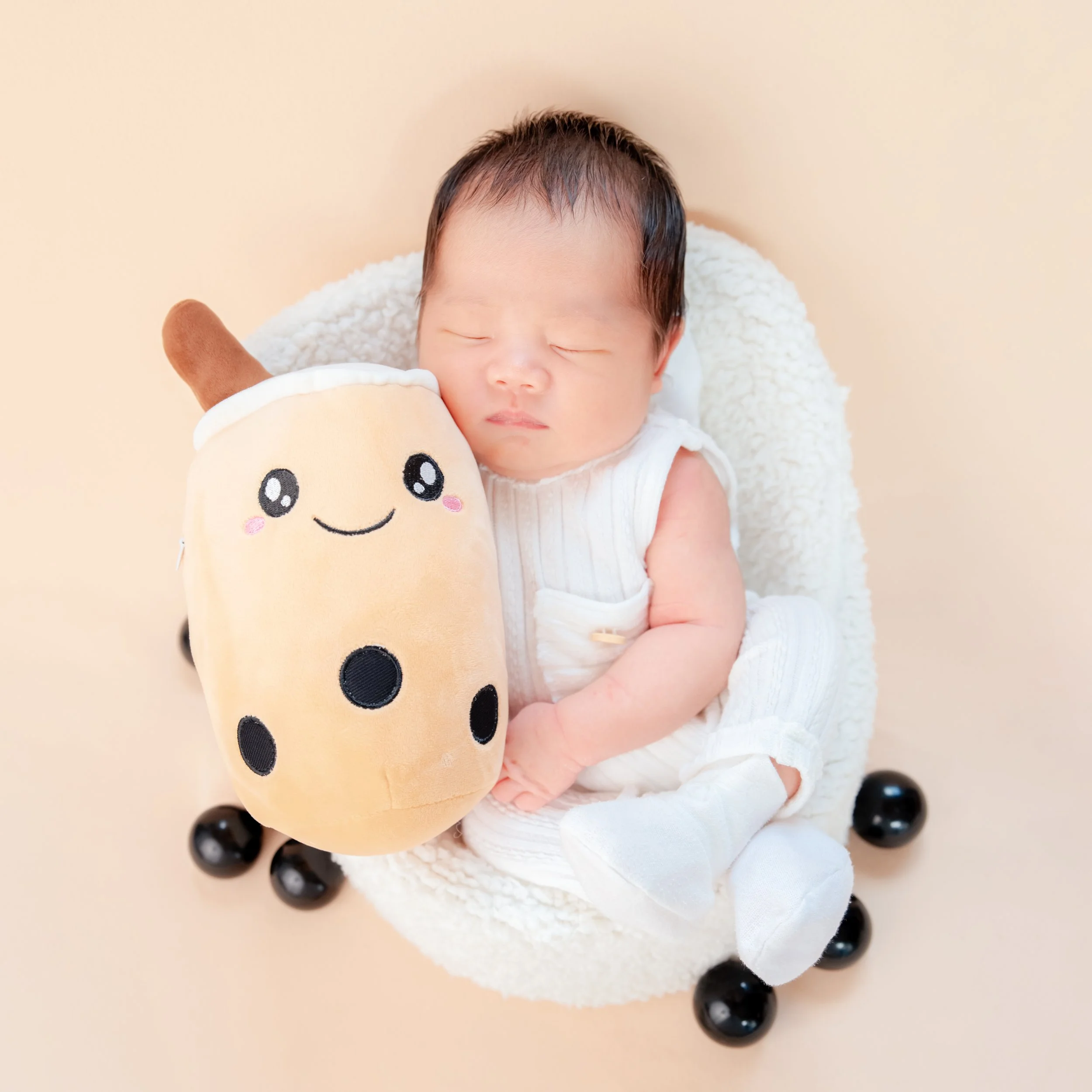 Sleeping newborn in a white knitted outfit cuddling a smiling bubble-tea plush in a white basket, with black pearls around