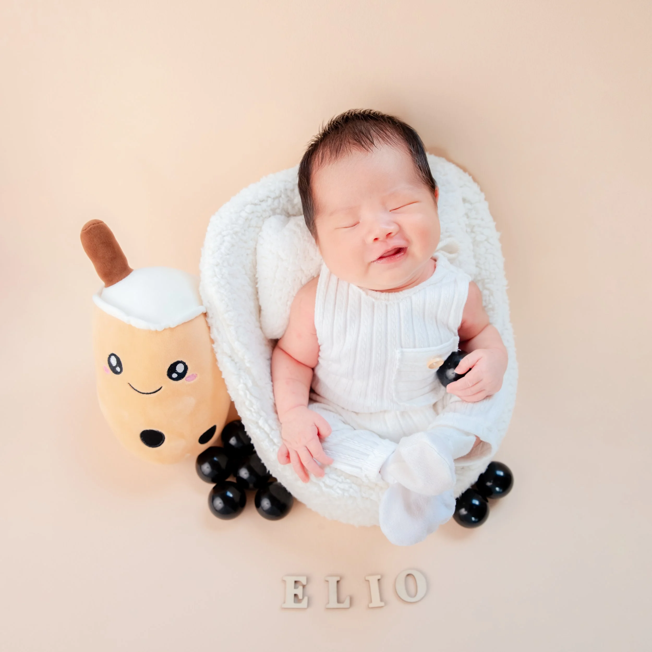 Newborn in a white knitted outfit lying in a white basket beside a smiling bubble-tea plush and black pearls