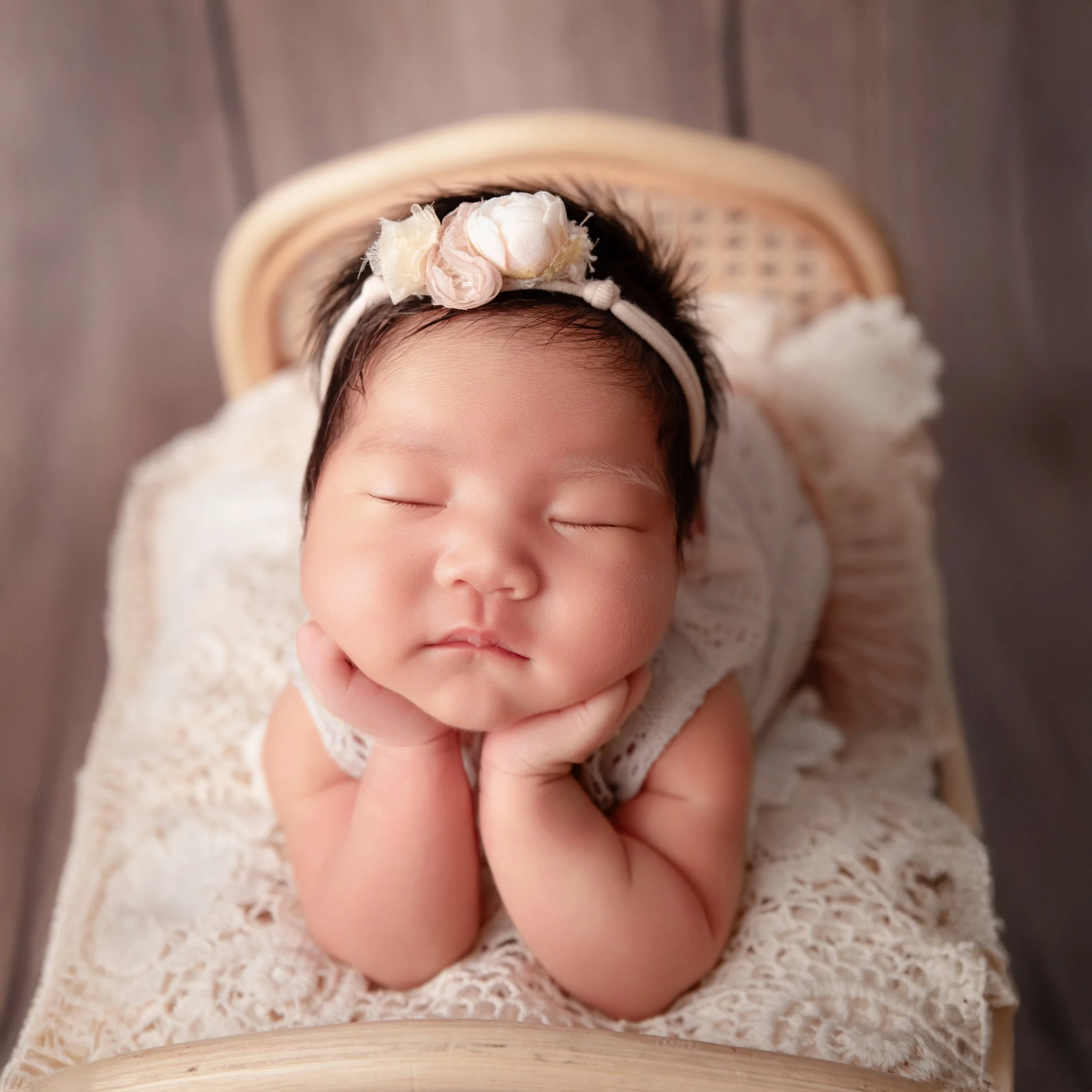 Close-up of a smiling sleeping newborn wearing a pink floral headband with cheeks resting on both hands on a cream lace blanket